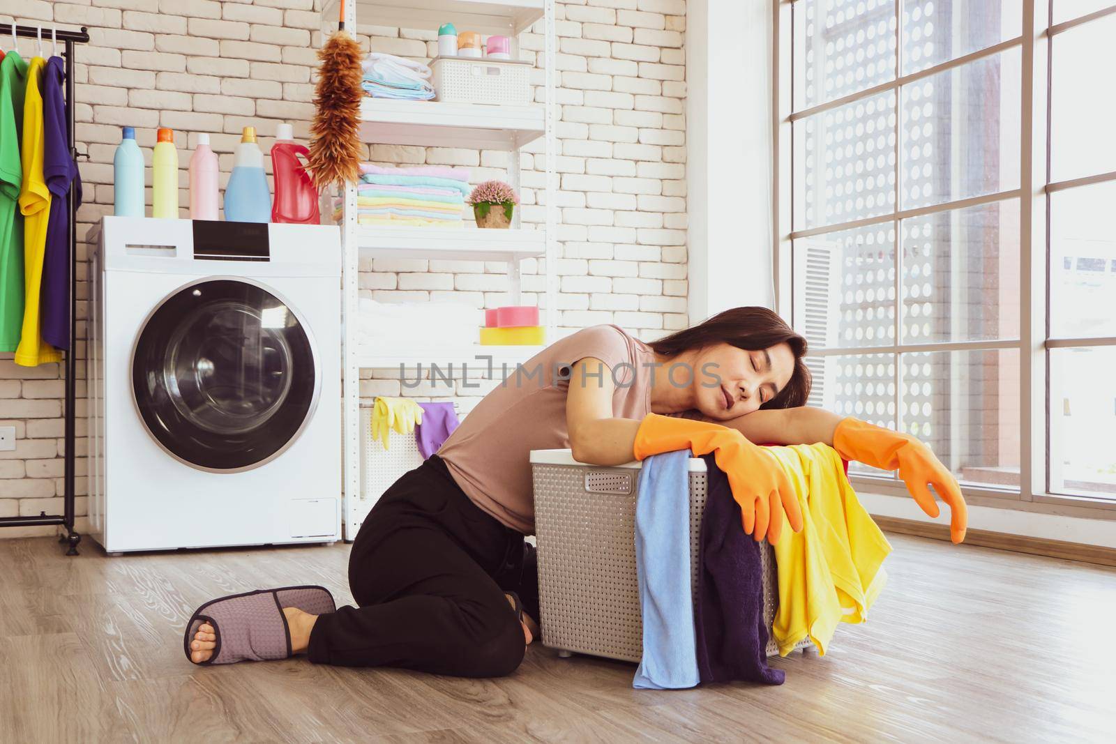 A cute Asian woman wearing a brown shirt. She is sleeping beside a laundry basket in the laundry room due to chores from doing housework and washing clothes during the day.