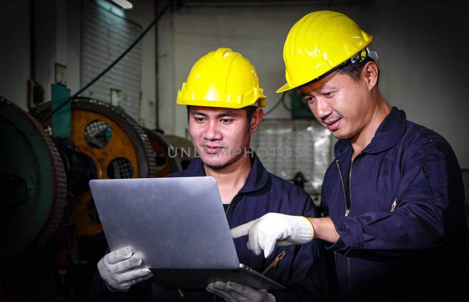 Two skilled Asian workers, dressed in helmet uniforms, are using a computer to analyze the metal press maintenance inspection data. The concept of teamwork at the factory