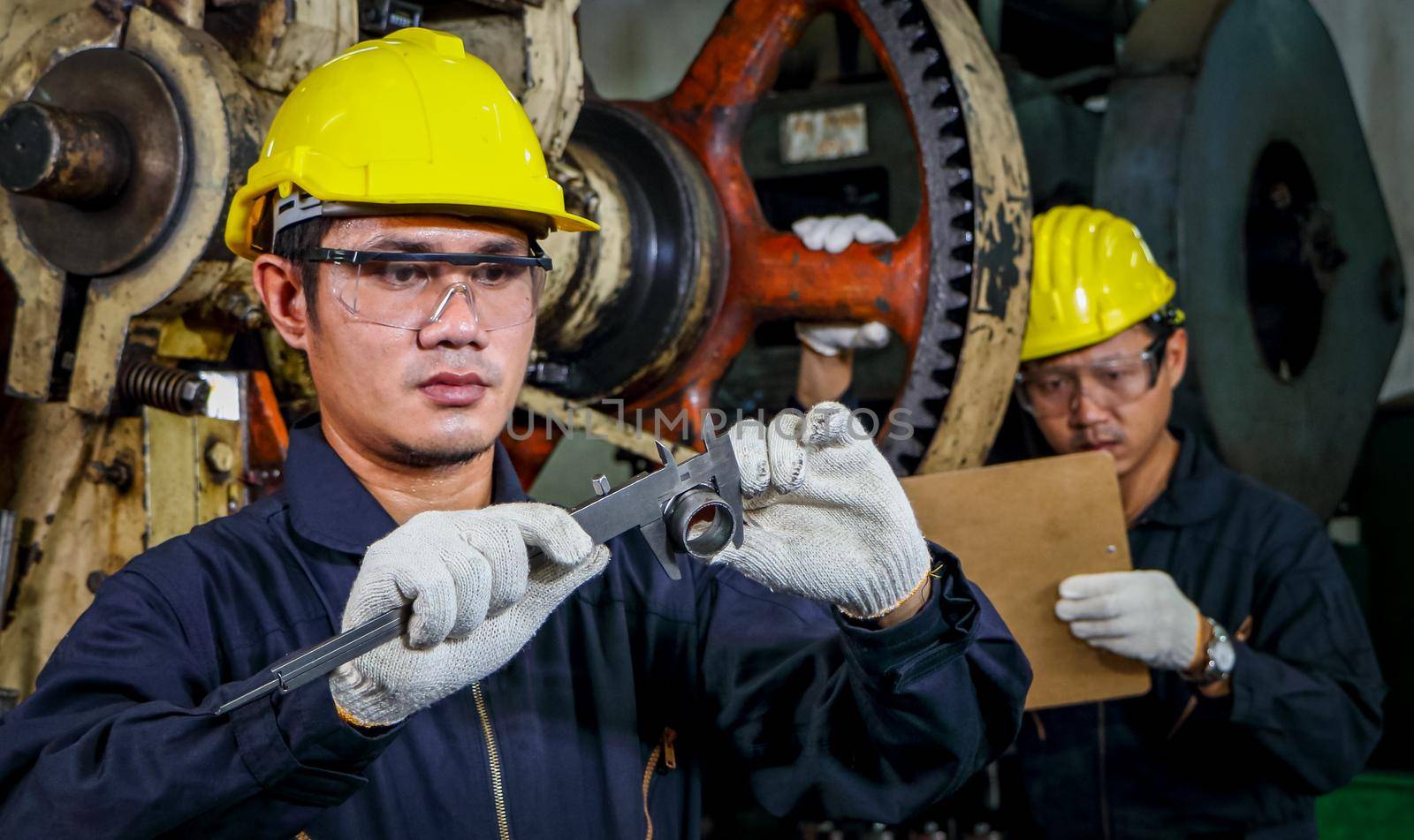 Two Asian male workers in safety clothing use Vernier calipers. Inspect the quality and measure the spare parts of working machines in industrial plants and production processes.