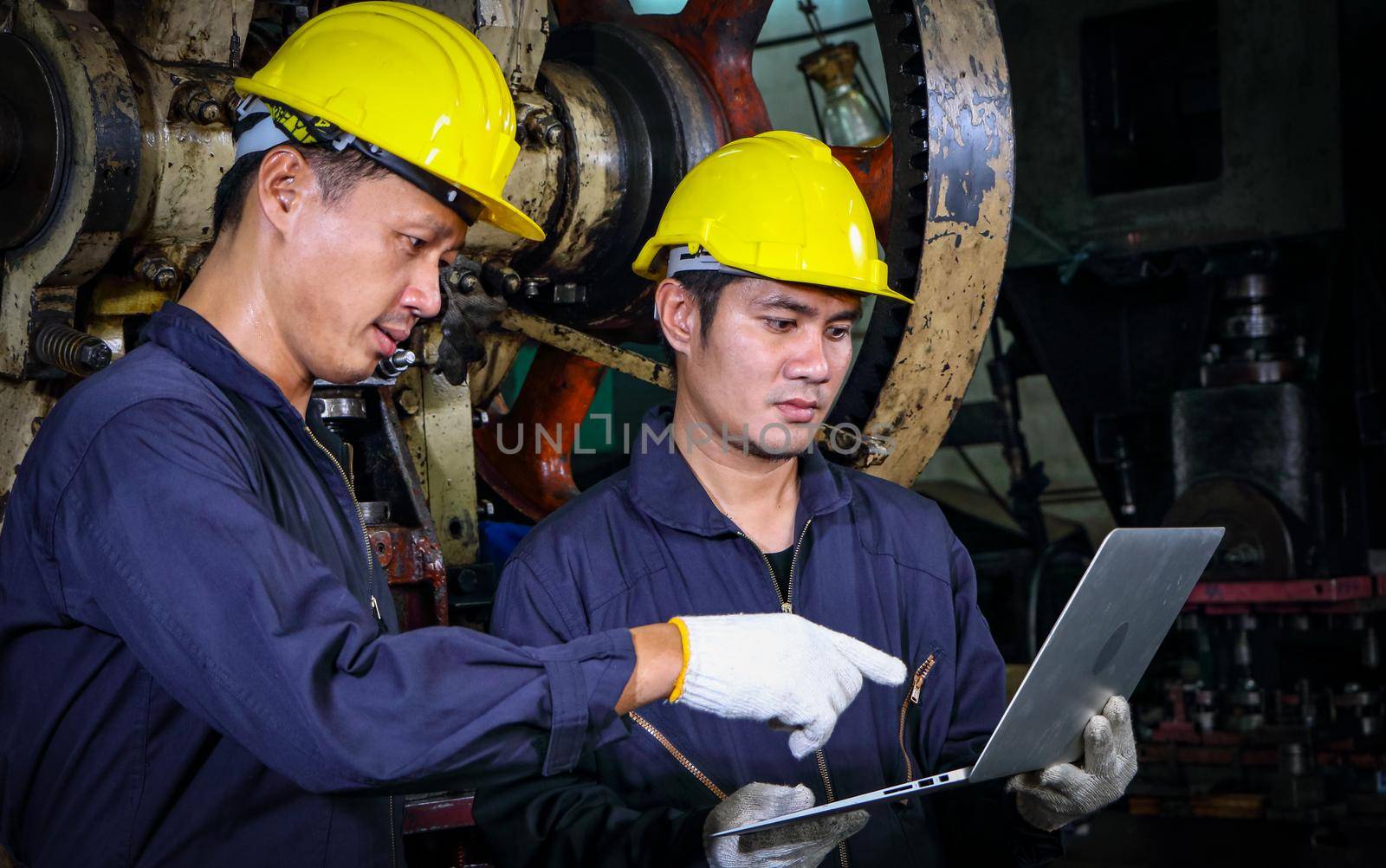 Two skilled Asian workers, dressed in helmet uniforms, are using a computer to analyze the metal press maintenance inspection data. The concept of teamwork at the factory