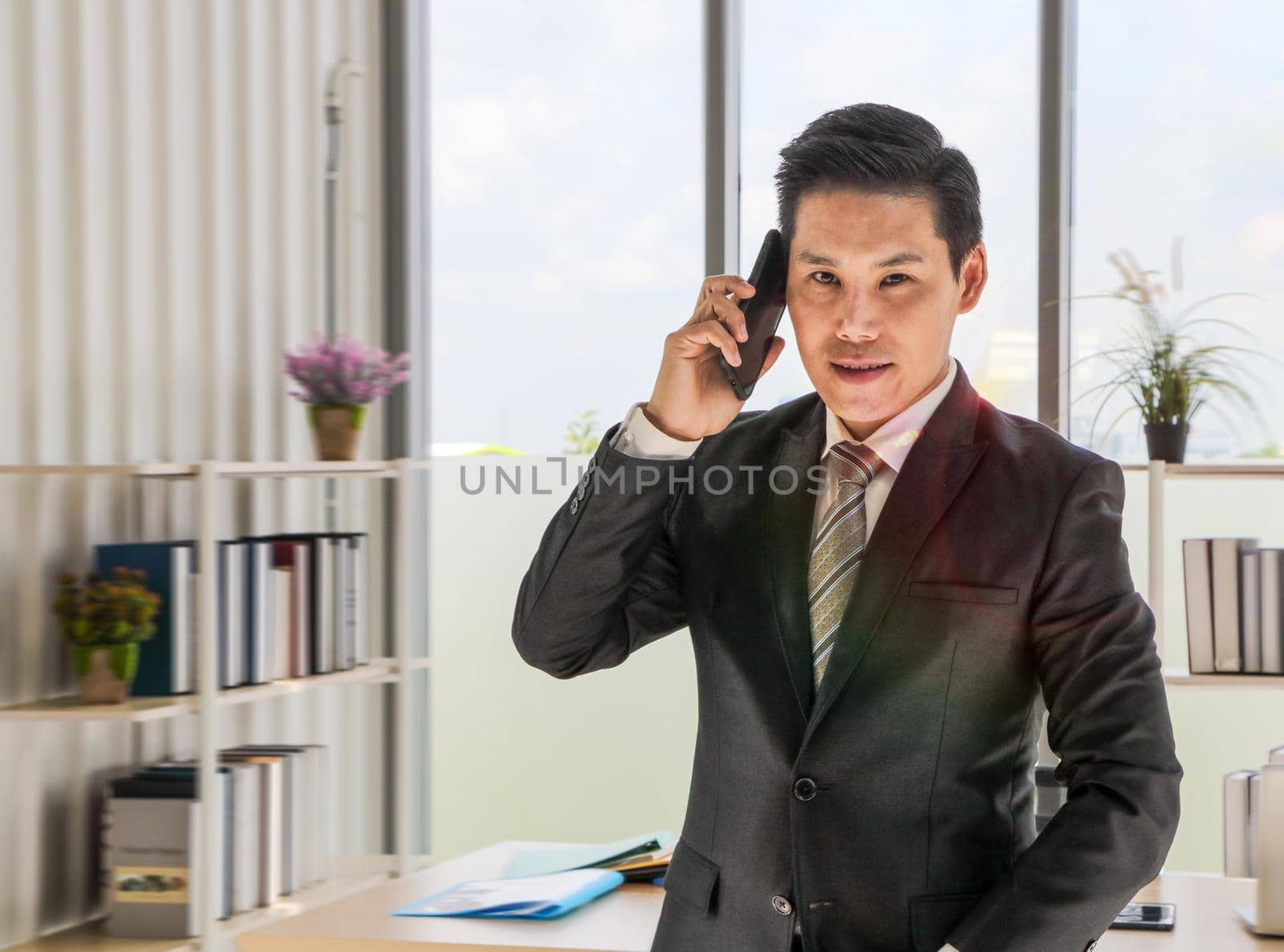 A young businessman in a black suit Who was happy to smile while using his smartphone to talk through the smartphone at the office desk in the sun shining down Business concept and copy space