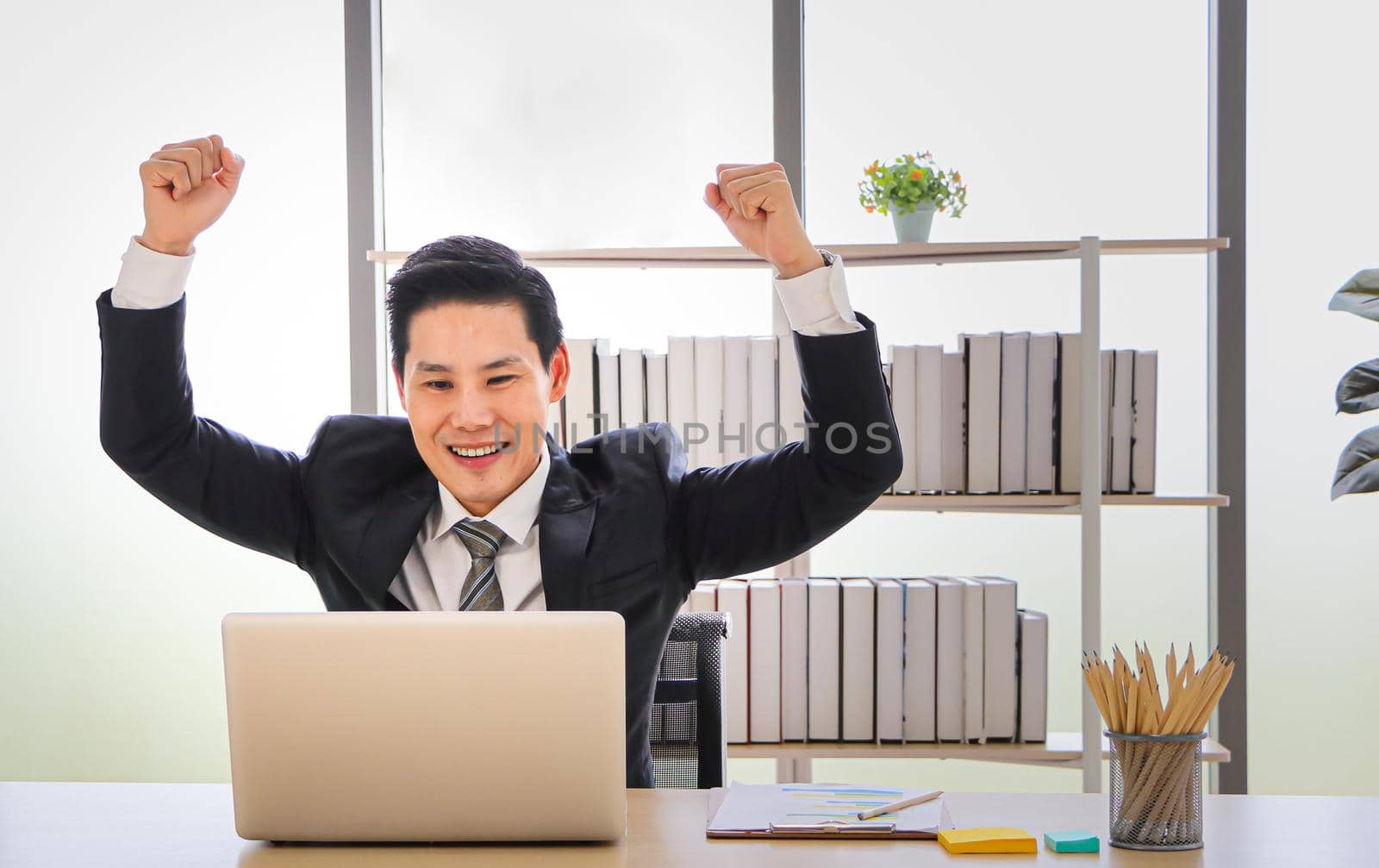 Portrait of an Asian businessman sitting at a table, looking at the PC screen, smiling and raising his hands on both sides, showing the joy of receiving an amazing fine rice celebrating success.