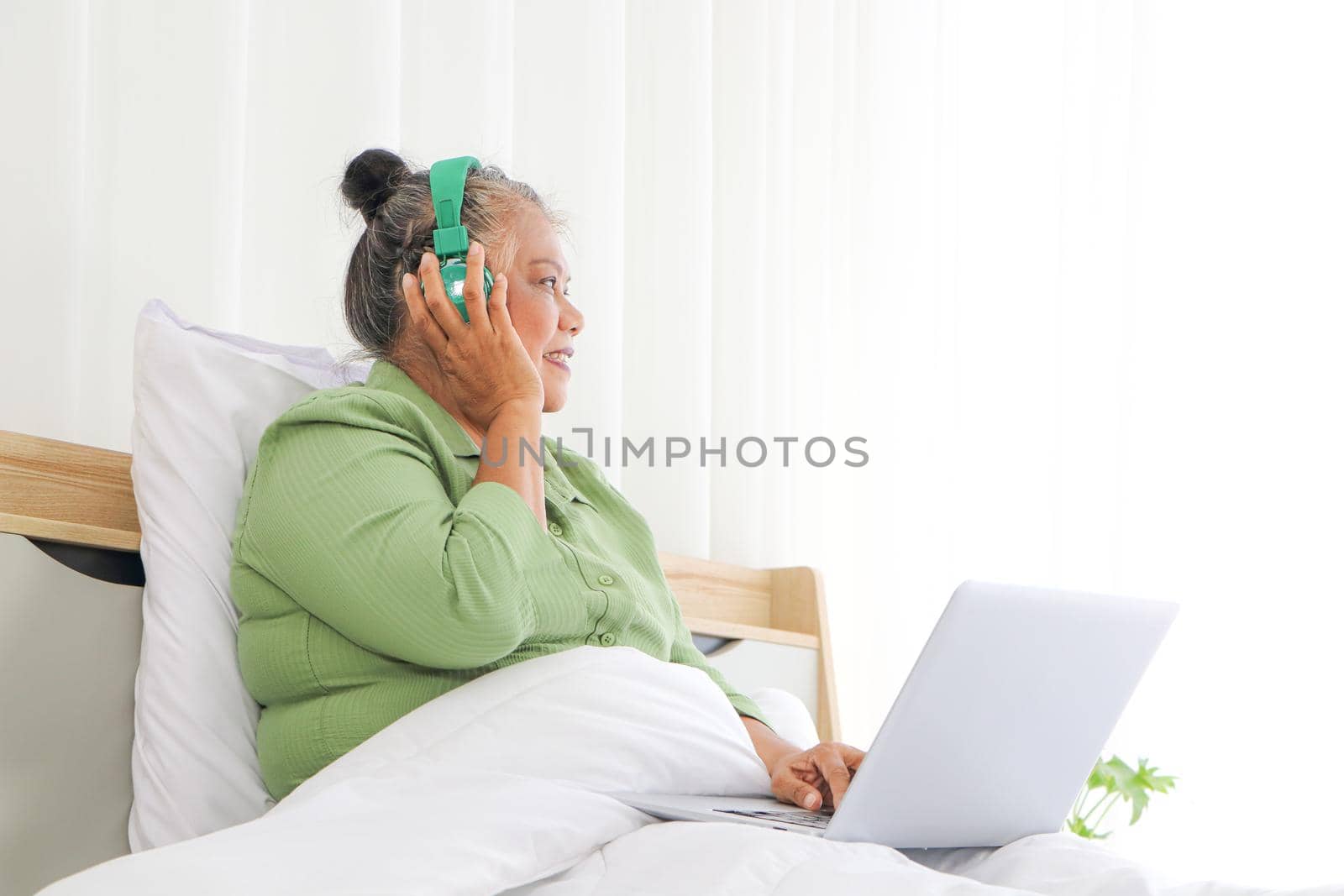 Retirement women Asians, use a laptop and headphones, happy in bed, concept listening to music, and living at home. Long-distance conversation through the online system Use of technology