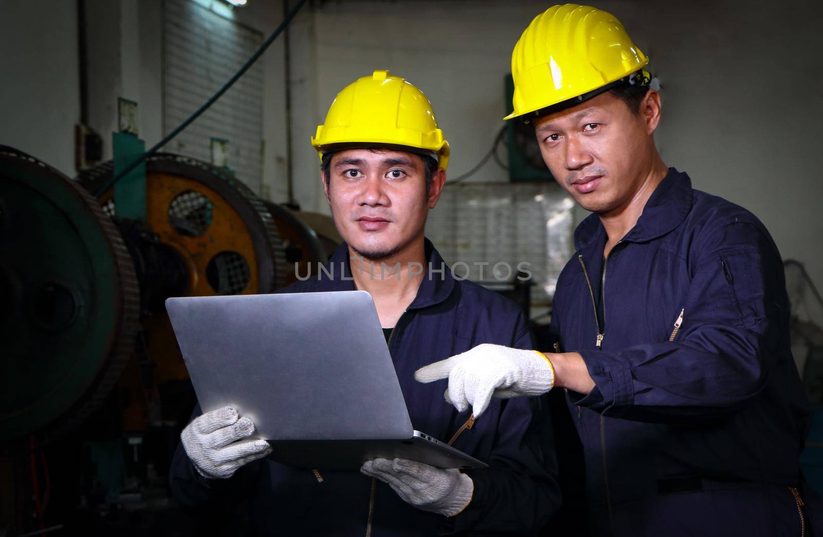 Two skilled Asian workers, dressed in helmet uniforms, are using a computer to analyze the metal press maintenance inspection data. The concept of teamwork at the factory