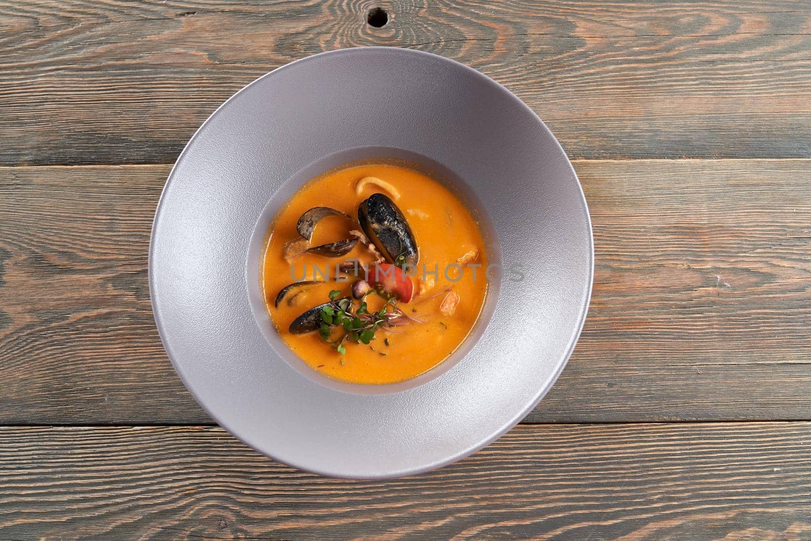 From above view of tasty bright orange soup with mussels in black shells in white plate on wooden restaurant table. Delicious seafood served with sprouts and tomatoes. Concept of exotic asian cuisine.