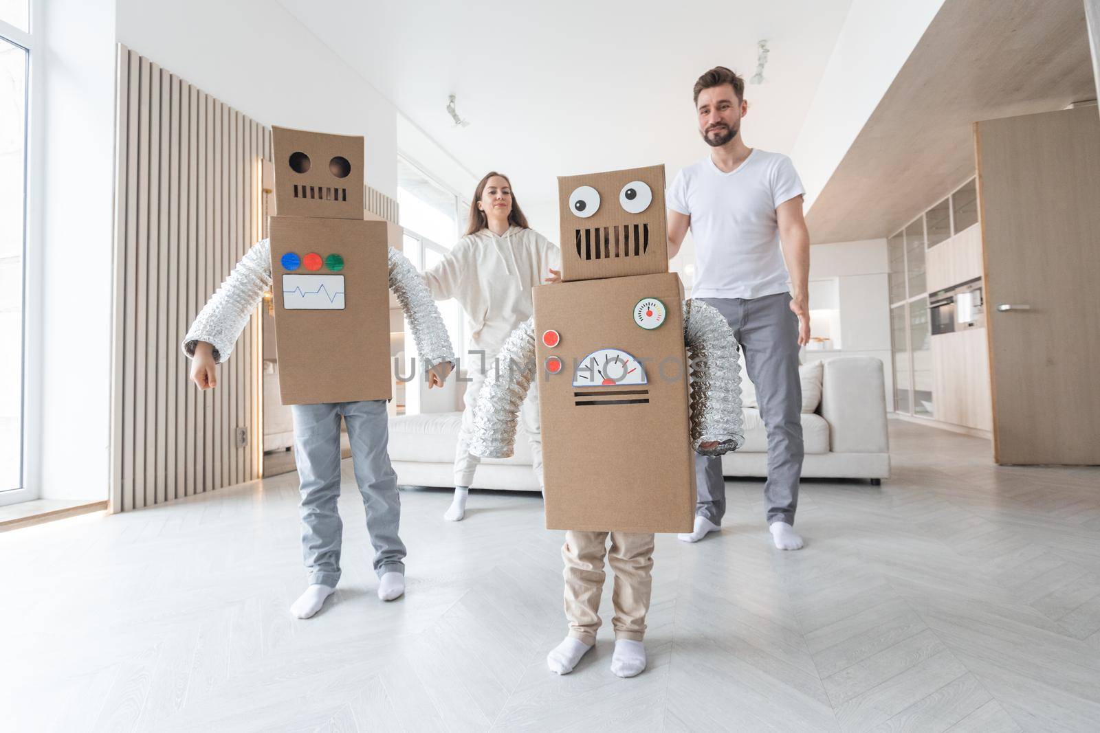 Happy family of parents and two children playing dancing like robots at home, children wearing handmade moving box costume of cardboard