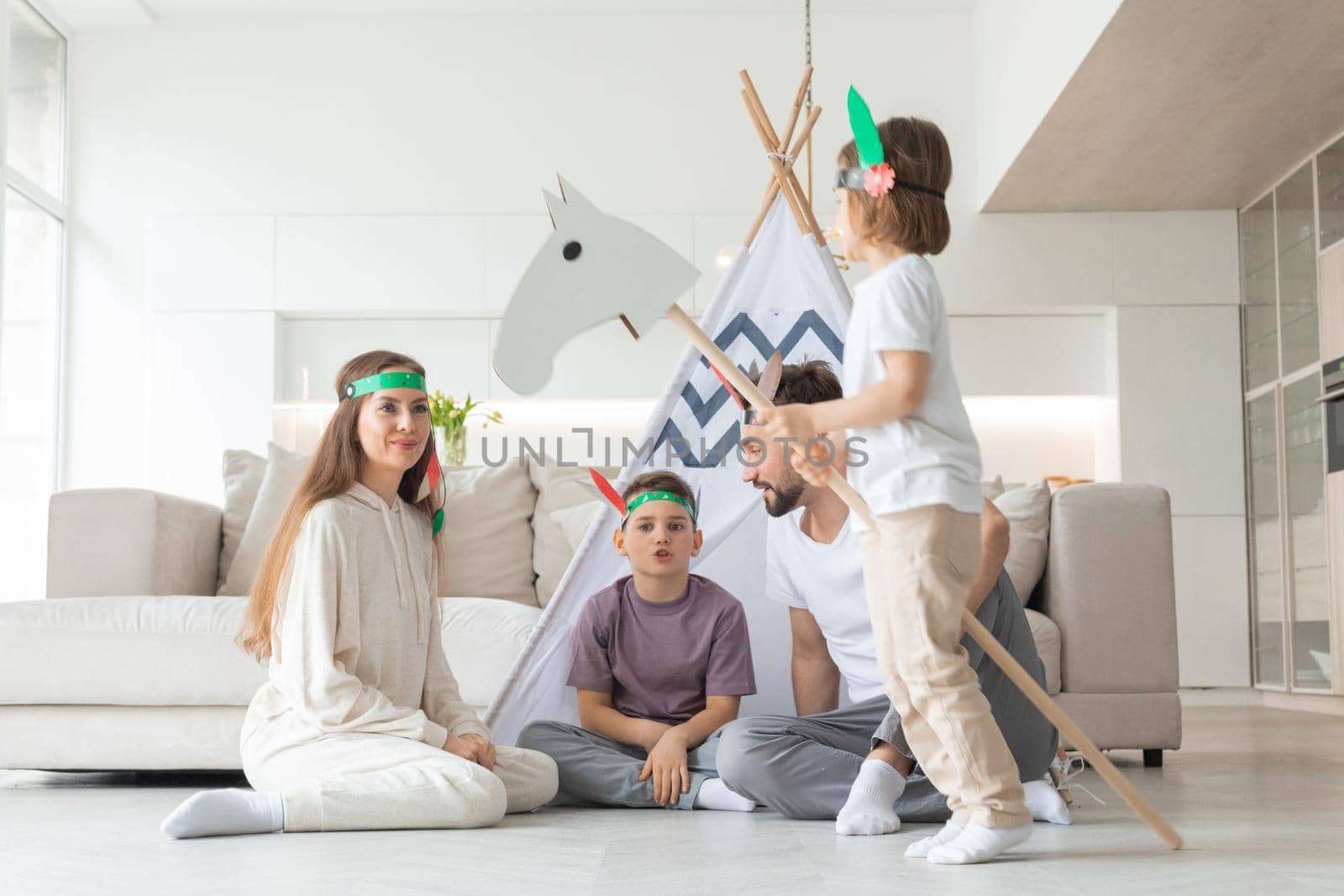 Happy family of parents and two children playing indian at home, wigwam tent, toy horse, feather roach