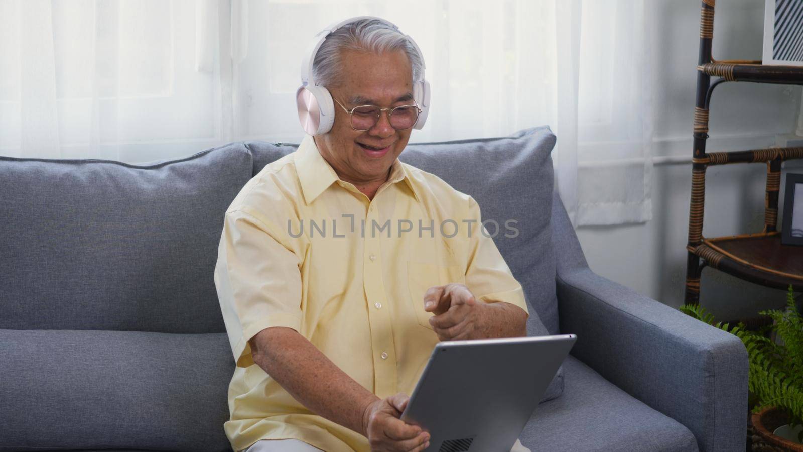 Old man grandfather smile with eyeglasses relaxing wear headphones is listening to music using a digital tablet, Asian happy senior old gray-haired sitting in living room on sofa at home video call