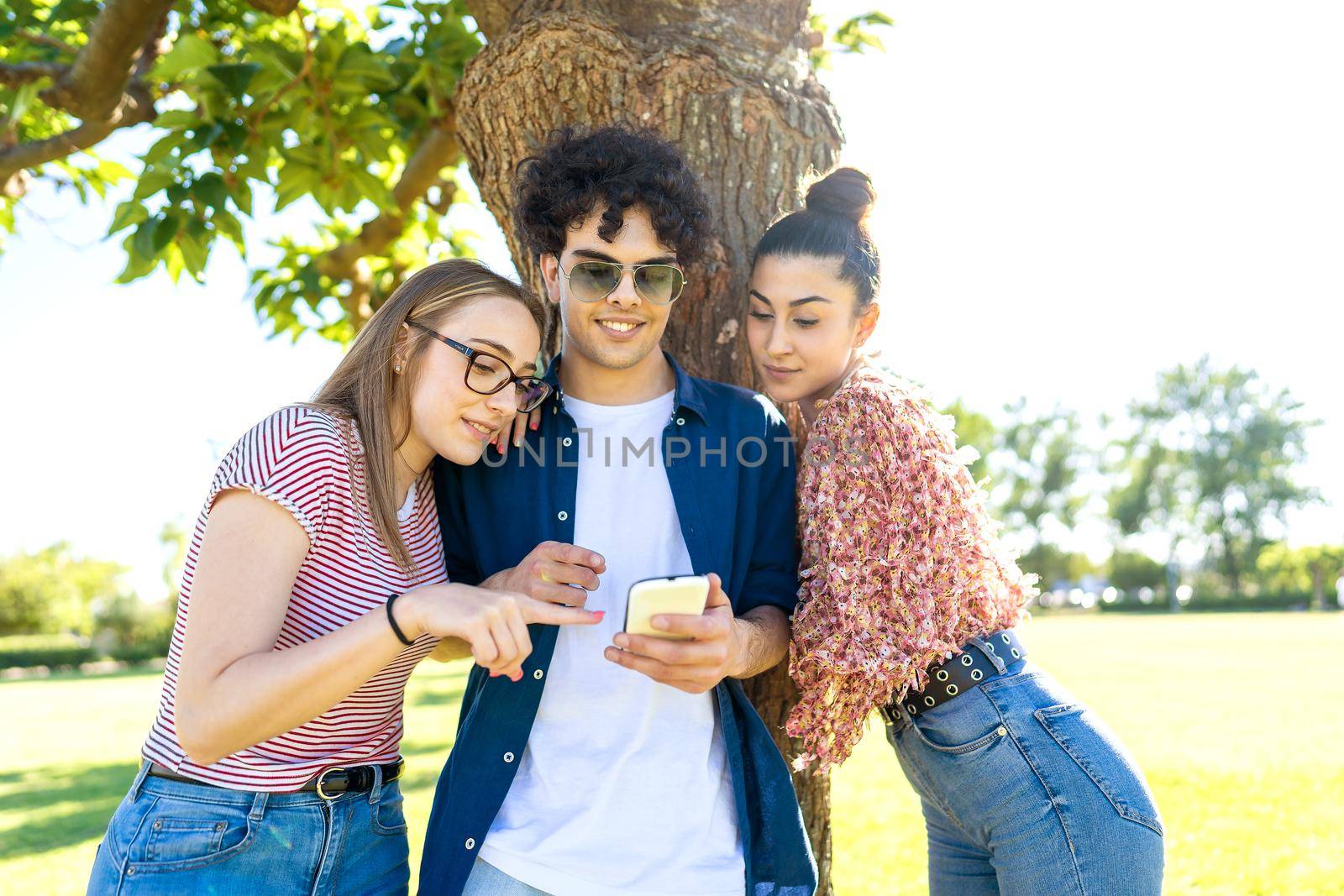 Three students having fun using smartphone at park pointing and texting on display. Two beautiful girls close to their best guy friend sharing on social network happy vacations moments