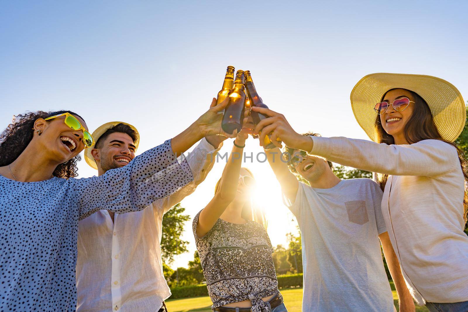 Group of young hipster friends celebrating outdoor drinking beer and toasting with bottle in front of sun setting. Happy beautiful millennials having fun with alcohol outdoor in a park at dusk by robbyfontanesi