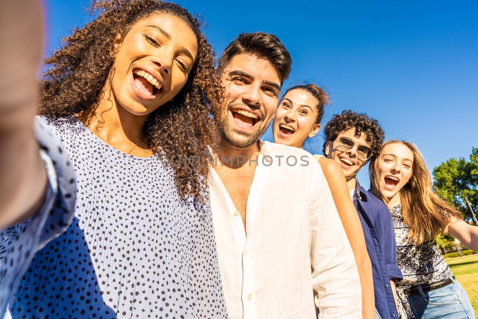 Five young multiracial friends in line posing for photo portrait smiling with open mouth. Black Hispanic brunette girl taking a selfie with smartphone outdoor. POV of happy millennial carefree people