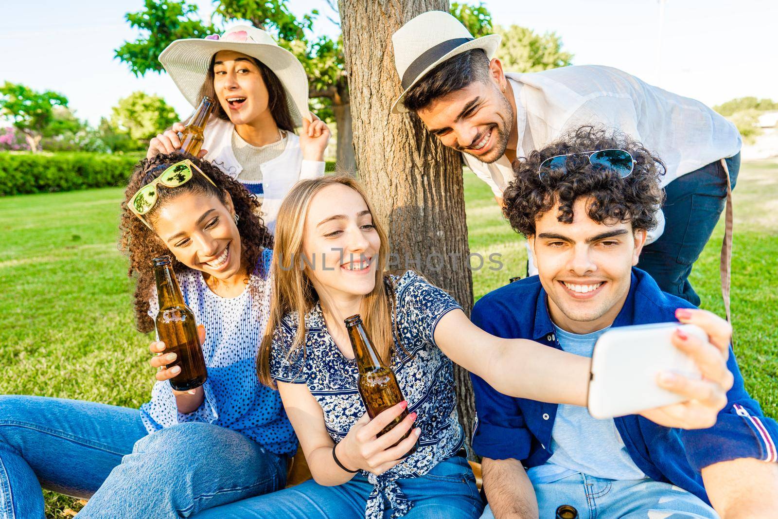 Five young multiracial friends having fun together in a picnic at city park drinking beer from bottle. Young blonde woman taking a self-portrait using smartphone to her multiracial diverse class mates by robbyfontanesi