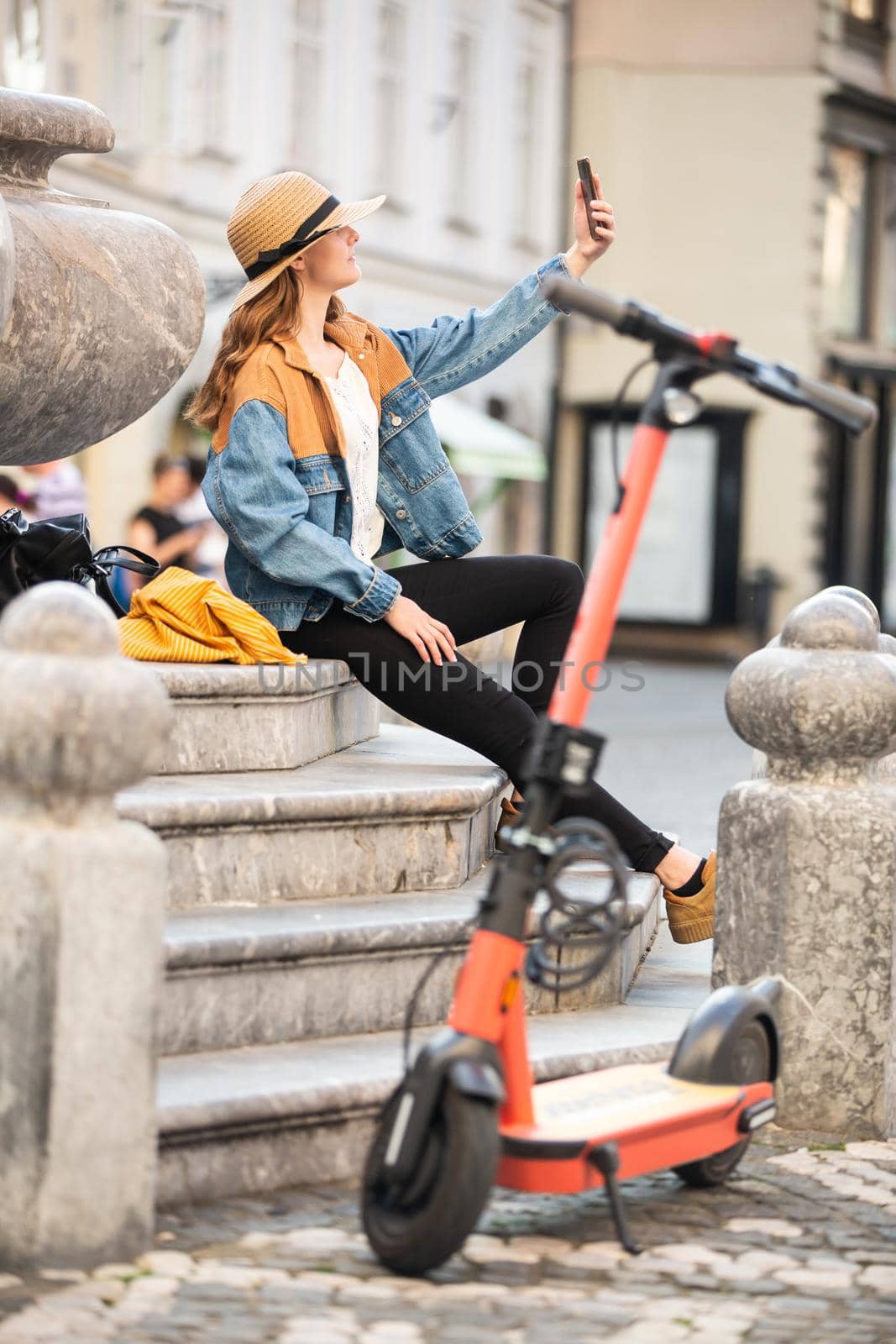 Young woman, sitting on staircase of old historical building in city center, taking picture with her phone. Female traveler exploring Ljubljana's old town on electric scooter. Summer leisure