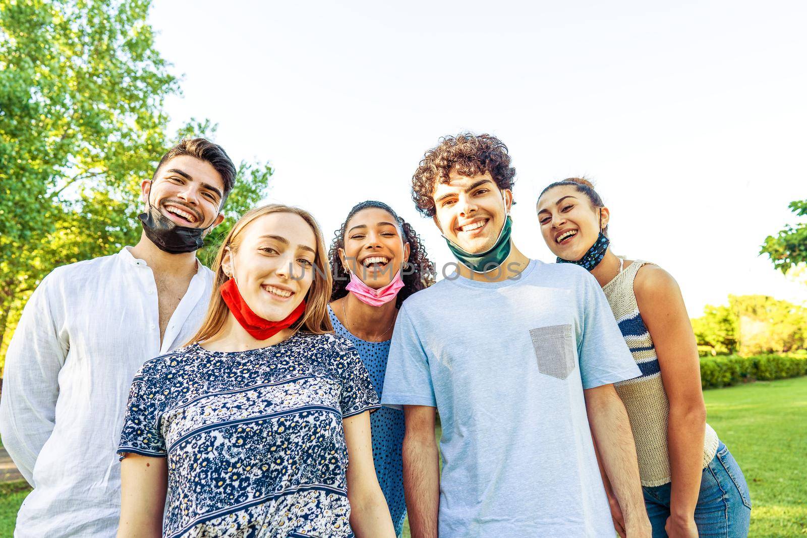Happy carefree group of multiracial friends posing in a park for a portrait wearing lowered protective face mask so showing their beautiful smiles. Concept of happiness despite difficulties of life