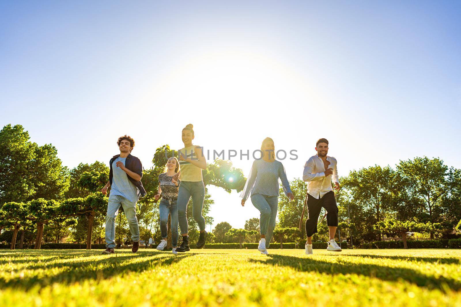 Suggestive low angle view of group of young multiracial gen-z friends running on green field with setting sun at their back with light effect and long shadows on grass. Concept of people team success