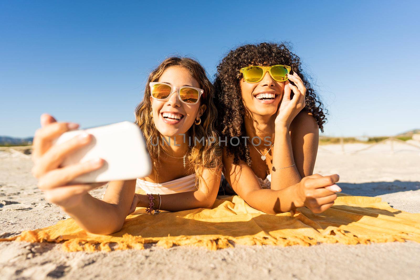Front portrait of two cute girls with sunglasses in summer vacation using smartphone taking a selfie lying on sea sand of a tropical beach. Multiracial women travellers having fun with technology by robbyfontanesi