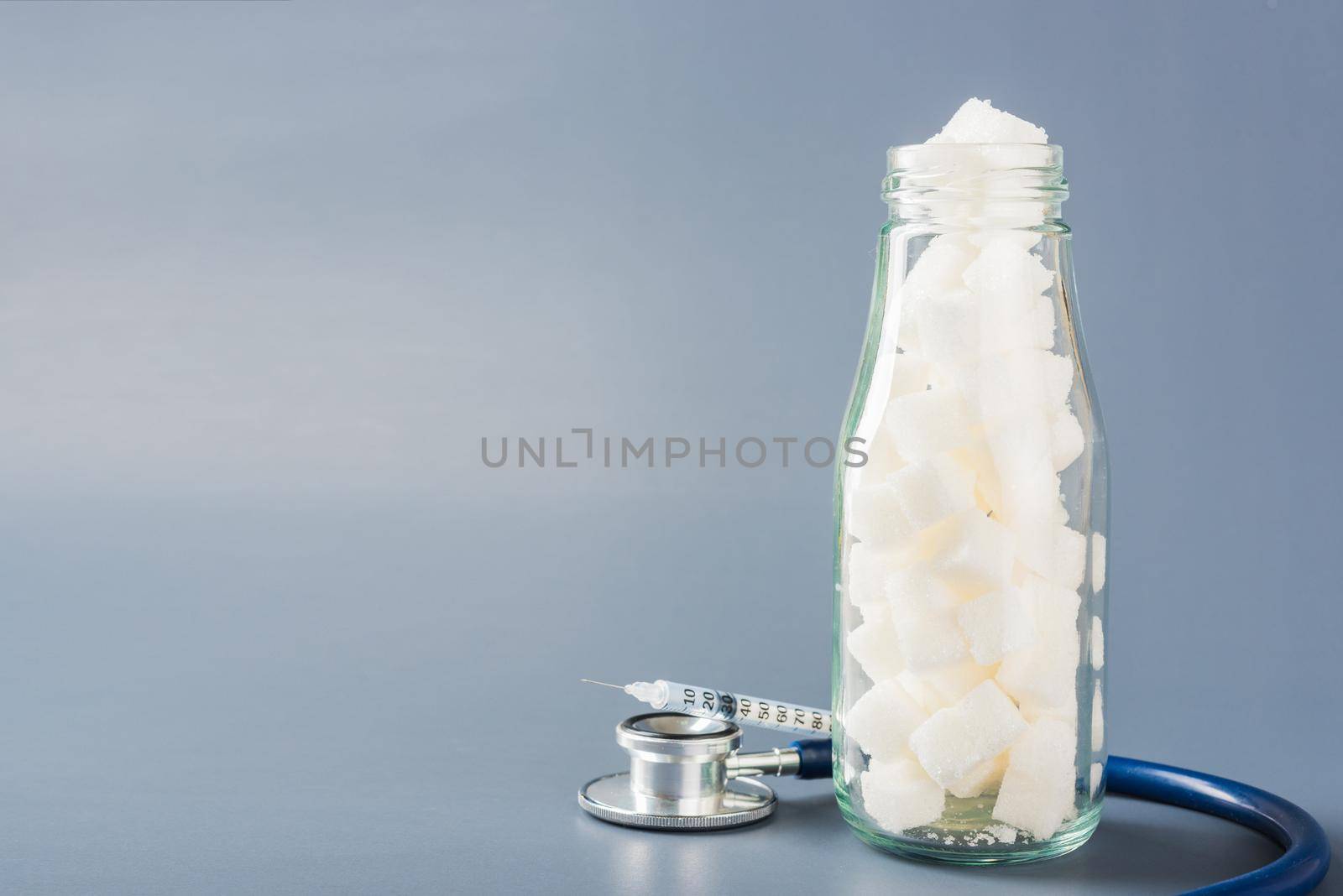 Glass bottle full of white sugar cube sweet food ingredient and doctor stethoscope isolated gray background, health high blood risk of diabetes and calorie intake concept and unhealthy drink