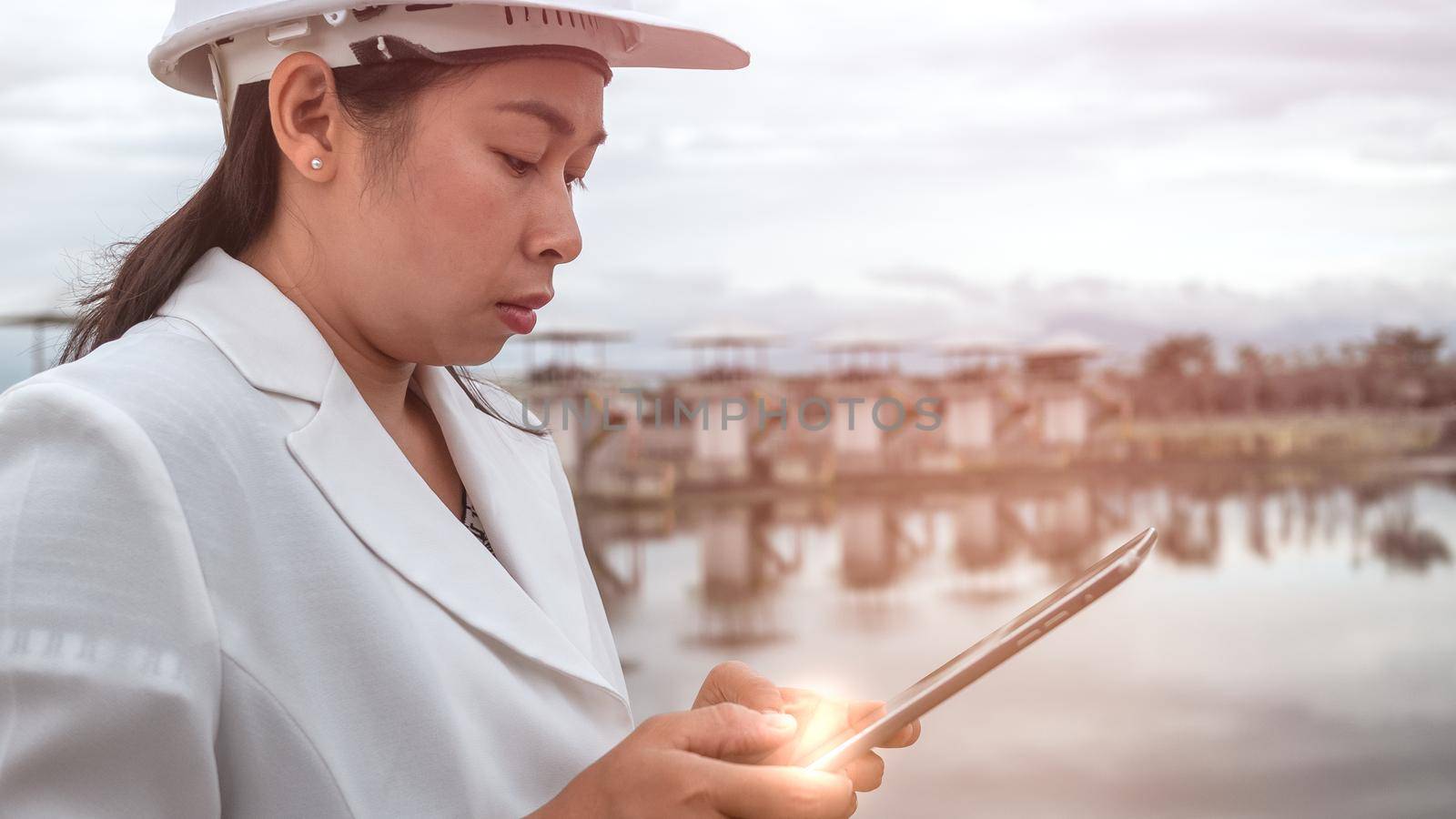 Female engineer in a white helmet reading and analyzing data in a digital tablet at the hydroelectric dam. Clean energy and Technology concepts. by TEERASAK