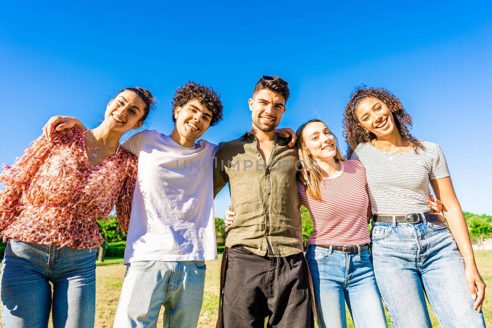 Multiracial group of college students embracing each other with arms around smiling looking at camera in a sunny day with blue sky at city park. Concept of equality and brotherhood among young people by robbyfontanesi