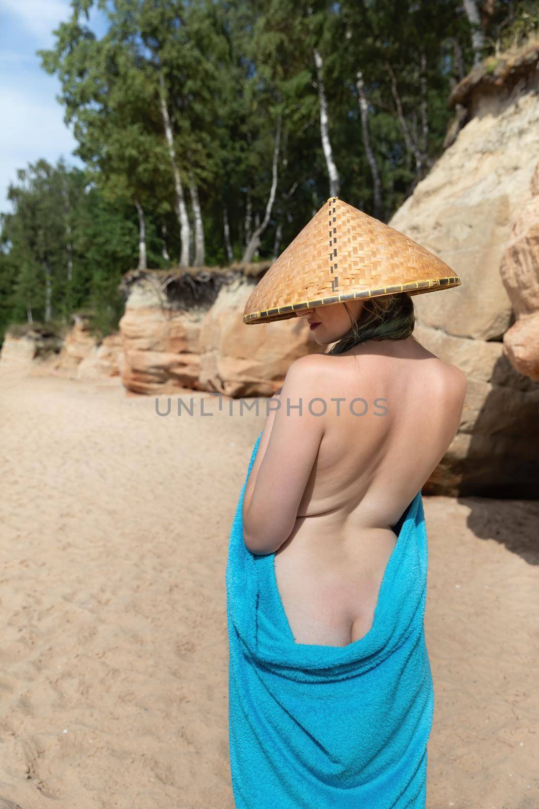 Young beautiful nude woman in straw vietnamese hat and a blue cape on the seashore near the sand caves