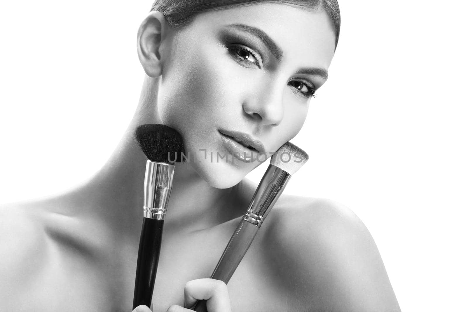 Glam beauty. Monochrome shot of a beautiful sensual young woman looking to the camera seductively holding makeup brushes to her face beauty cosmetic products maquillage style fashion concept