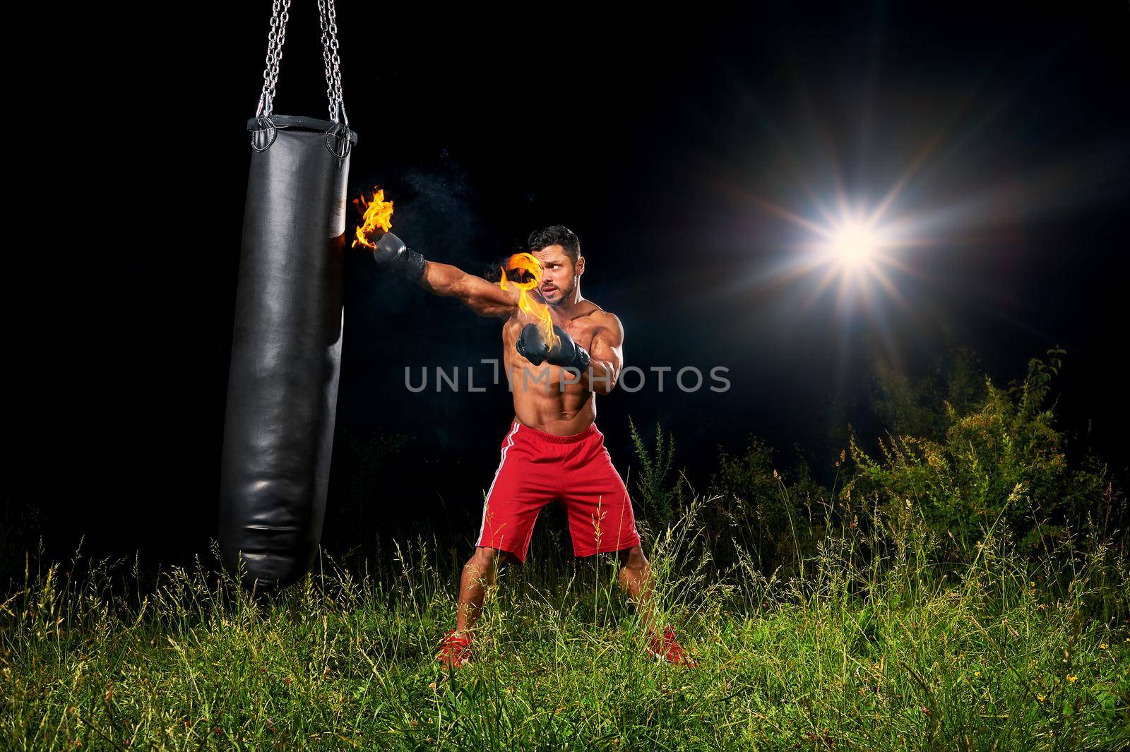 Shirtless muscular male boxer training outdoors at night exercising on a punching bag with his boxing gloves burning with fire copyspace fitness lifestyle forceful powerful confidence athlete.