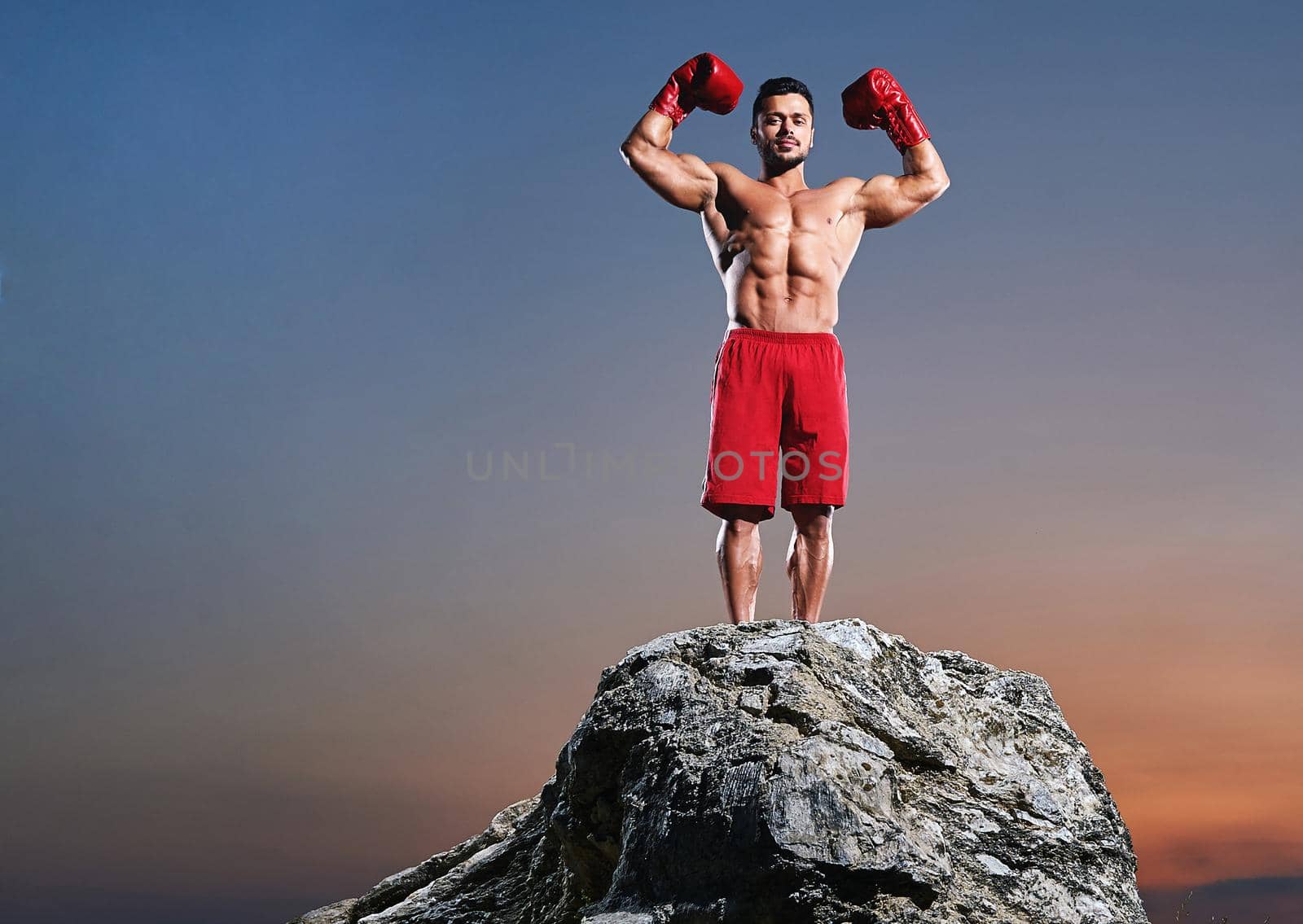 Horizontal full length shot of a strong muscular male boxer flexing his biceps standing on top of a rock outdoors on sunset copyspace champion winner fighter competition fitness sportsman sport .