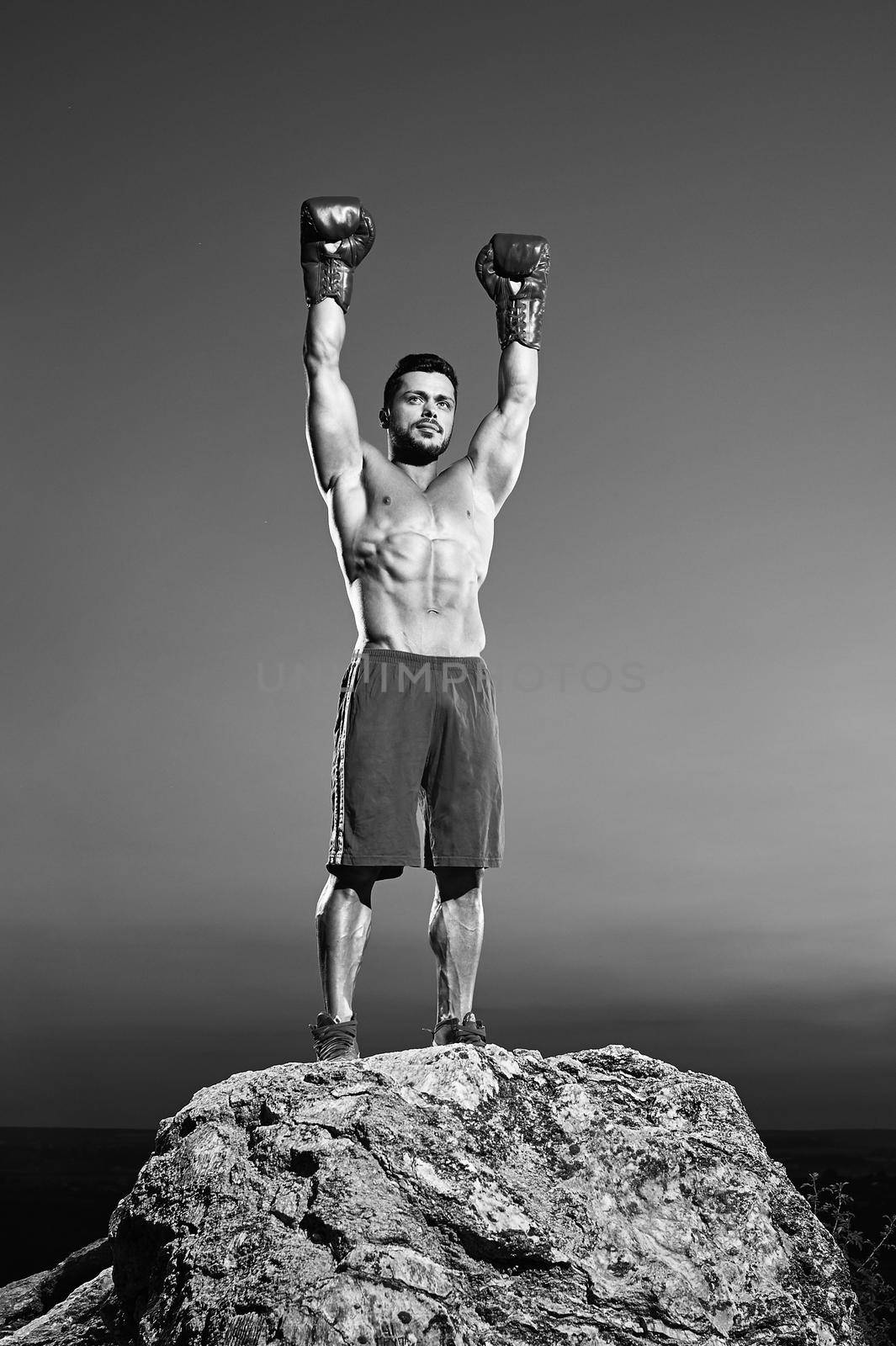 Full length vertical monochrome portrait of a boxing champion celebrating winning posing outdoors standing on top of a rock raising his arms in victory lifestyle achieving winner sports motivation.