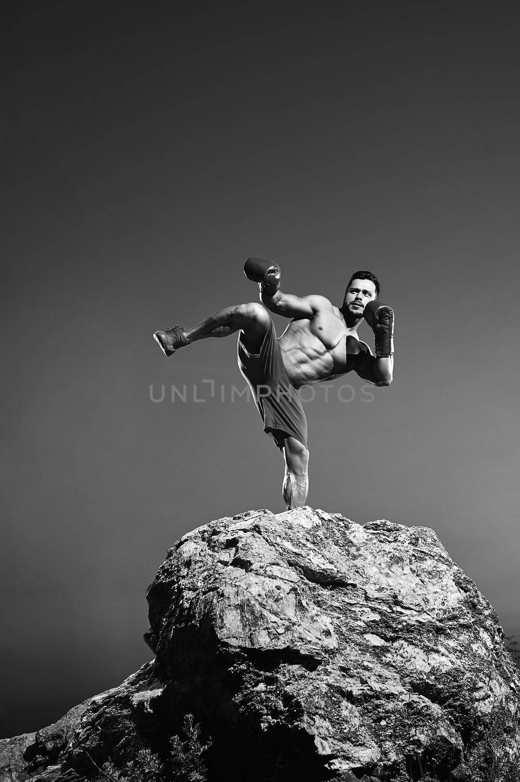 Black and white portrait of a male fighter training outdoors performing kickboxing technique boxing kicking strength balance martial combat fighting masculinity abs fitness sportive athlete physique.