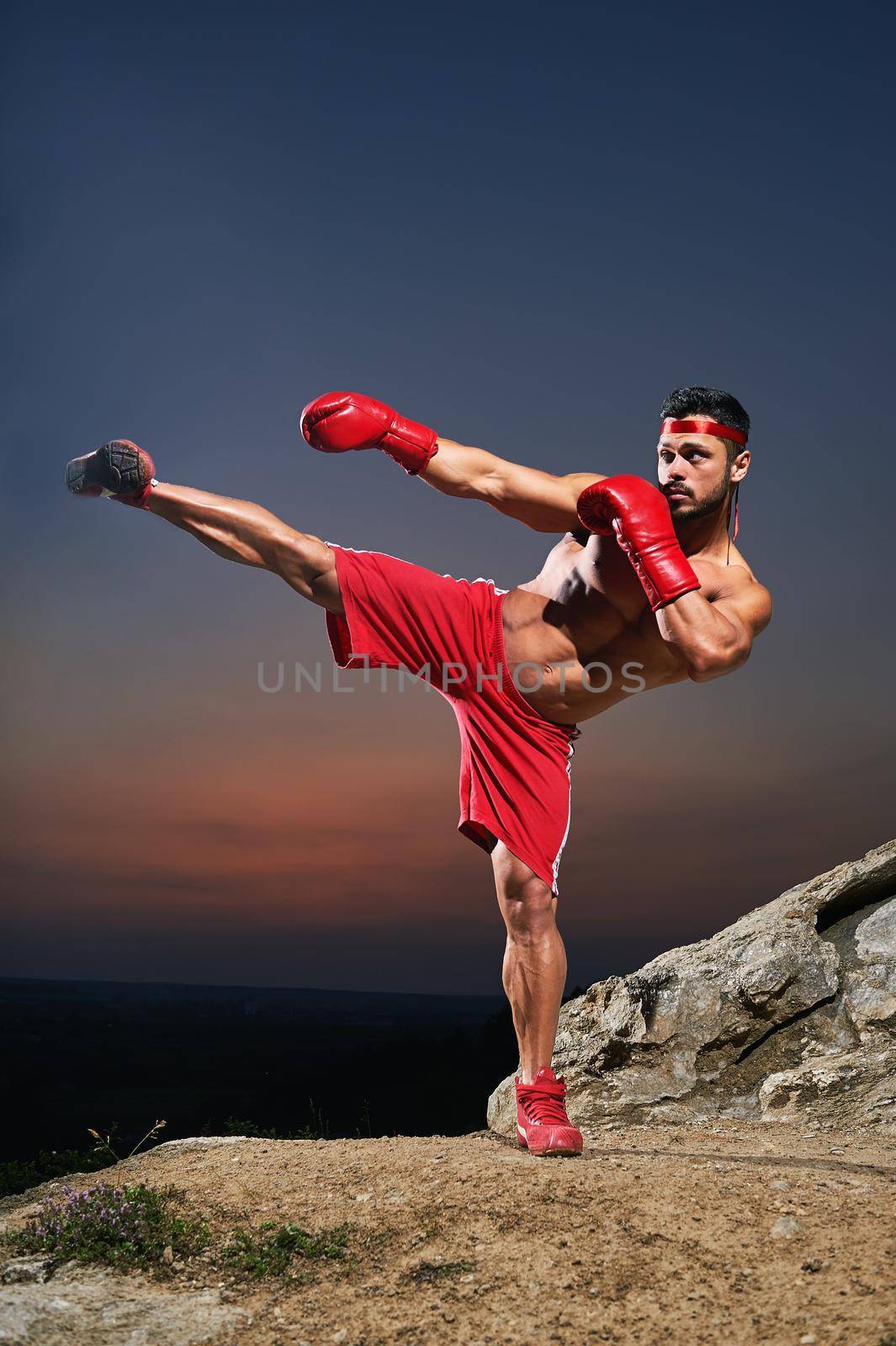 Vertical shot of a muscular strong male fighter practicing kickboxing training outdoors wearing boxing gloves sunset on the background sports motivation determination achievement strength combat