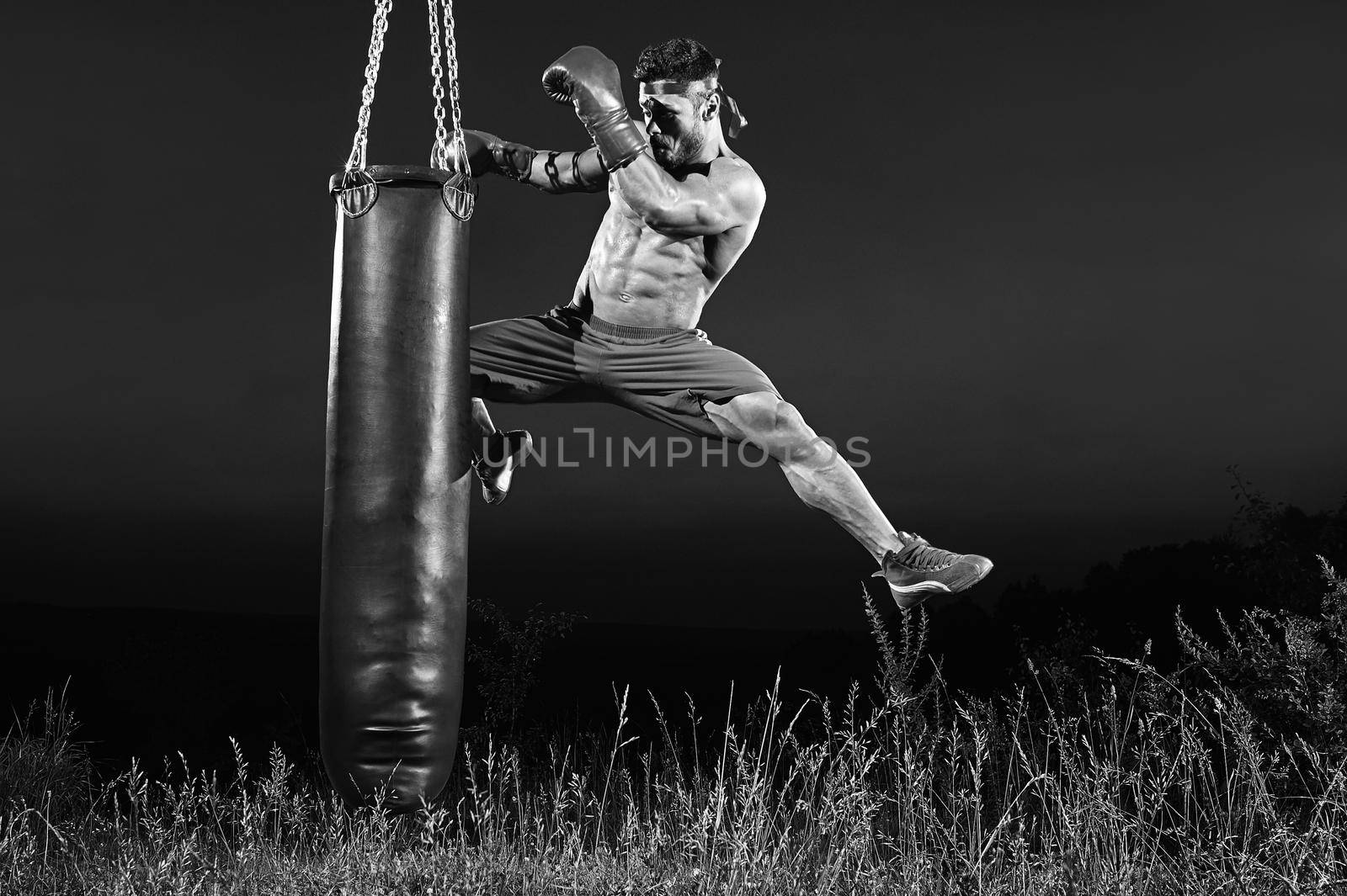 Black and white shot of a male kick boxer jumping and kicking a heavy punching bag training outdoors copyspace professional skilled motivation sports competitive preparing achievement fighting toned.