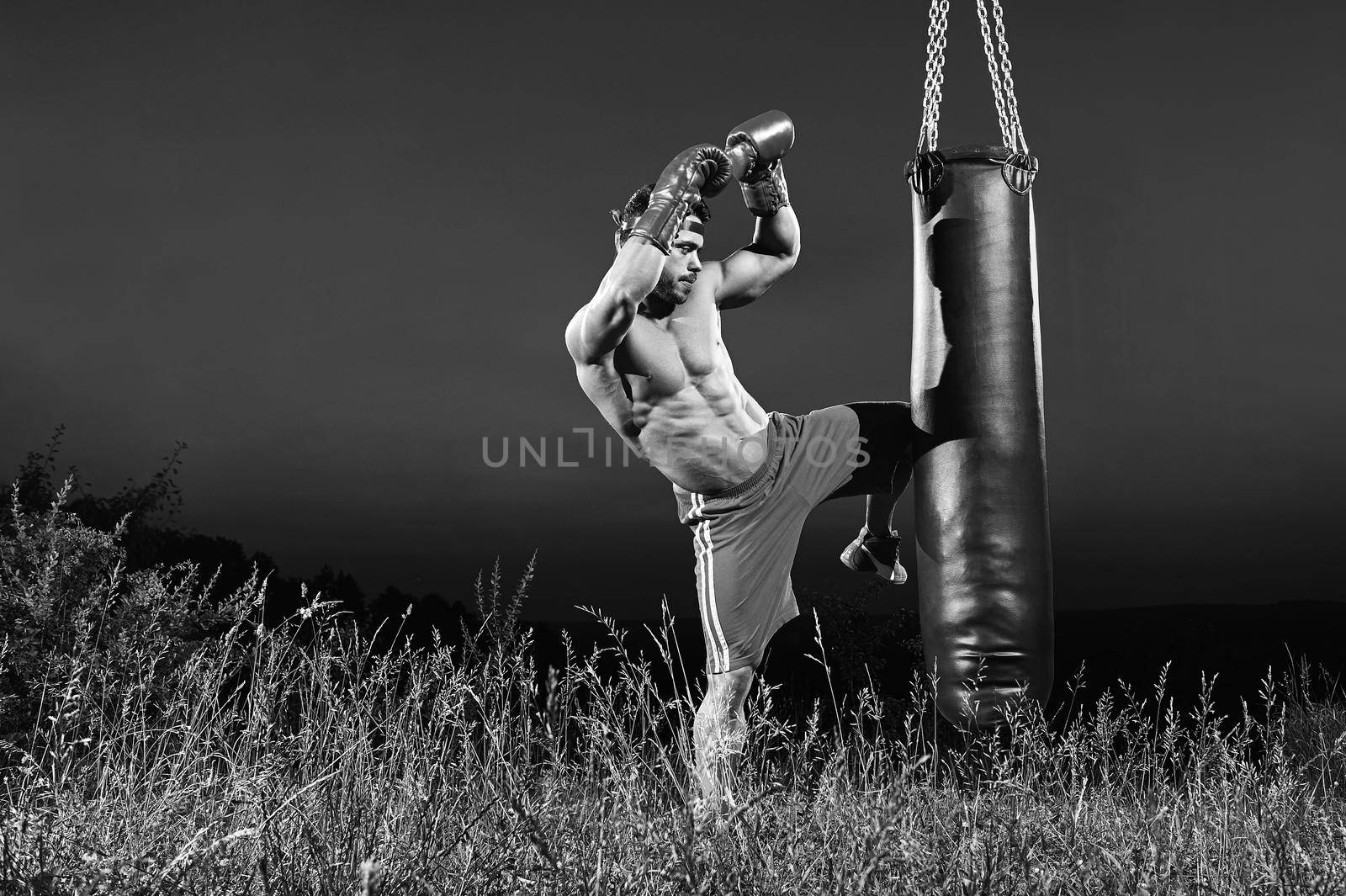 Black and white shot of a kick boxer training on a heavy bag outdoors kicking it with his knee copyspace kickboxing martial arts combat fighting practicing performance concentration determination.