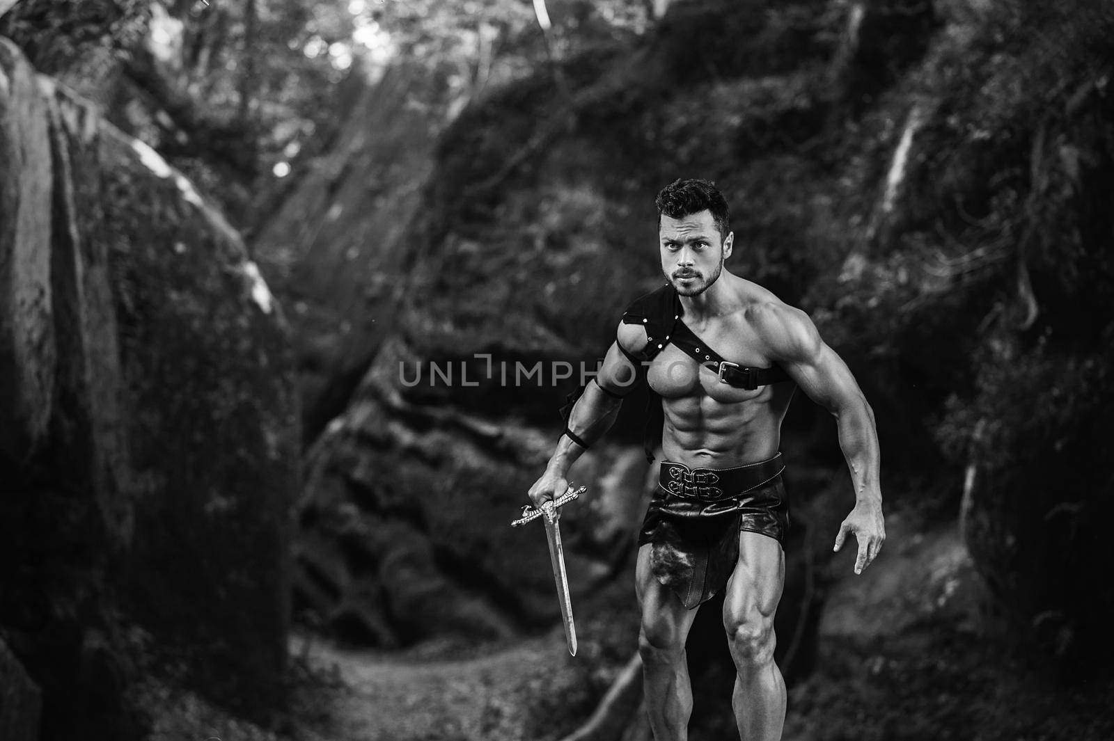 Come at me. Monochrome shot of a young manly gladiator holding a sword ready to fight near the rocks