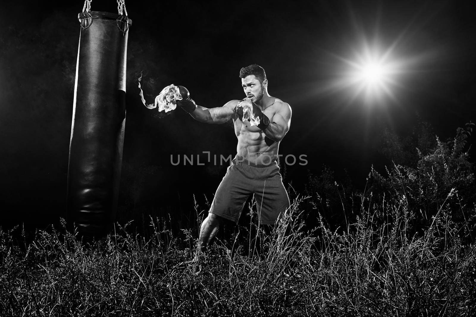 Monochrome shot of a boxer with flaming boxing gloves hitting a punching bag outdoors training at night copyspace sports motivation concentration focus burning fire burn fighting practicing workout.
