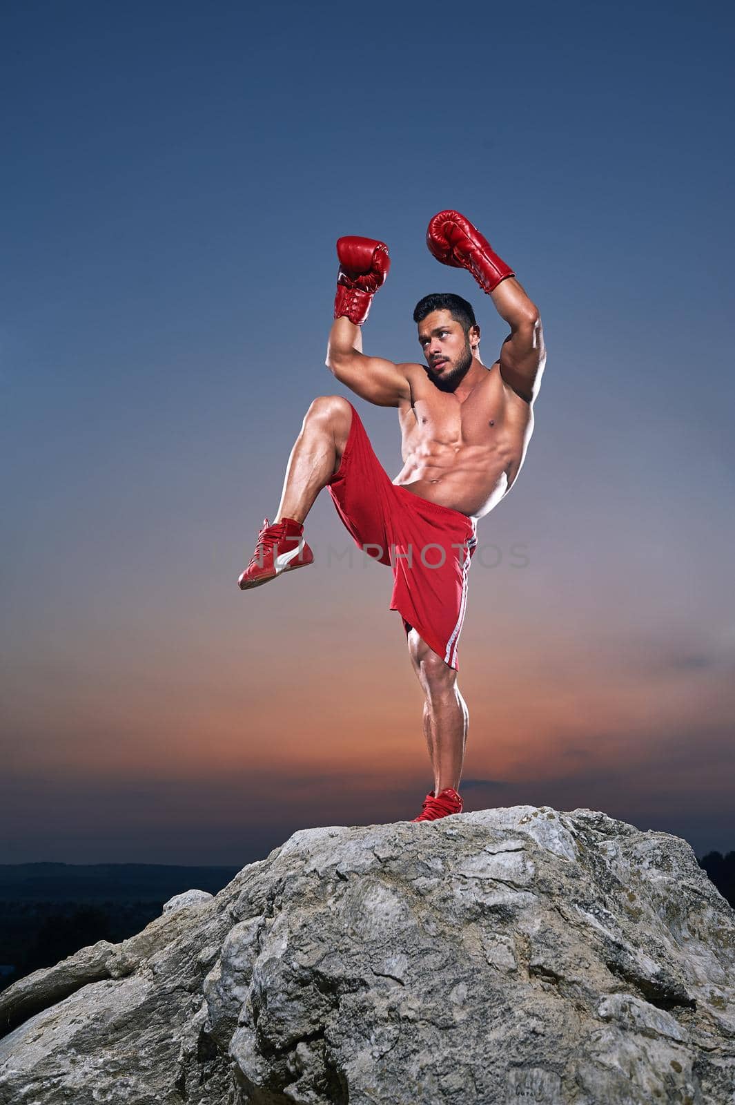 Vertical shot of a muscular young strong fighter wearing boxing gloves training outdoors doing kickboxing exercises shirtless muscular fighting torso abs ripped toned fit fitness health lifestyle.
