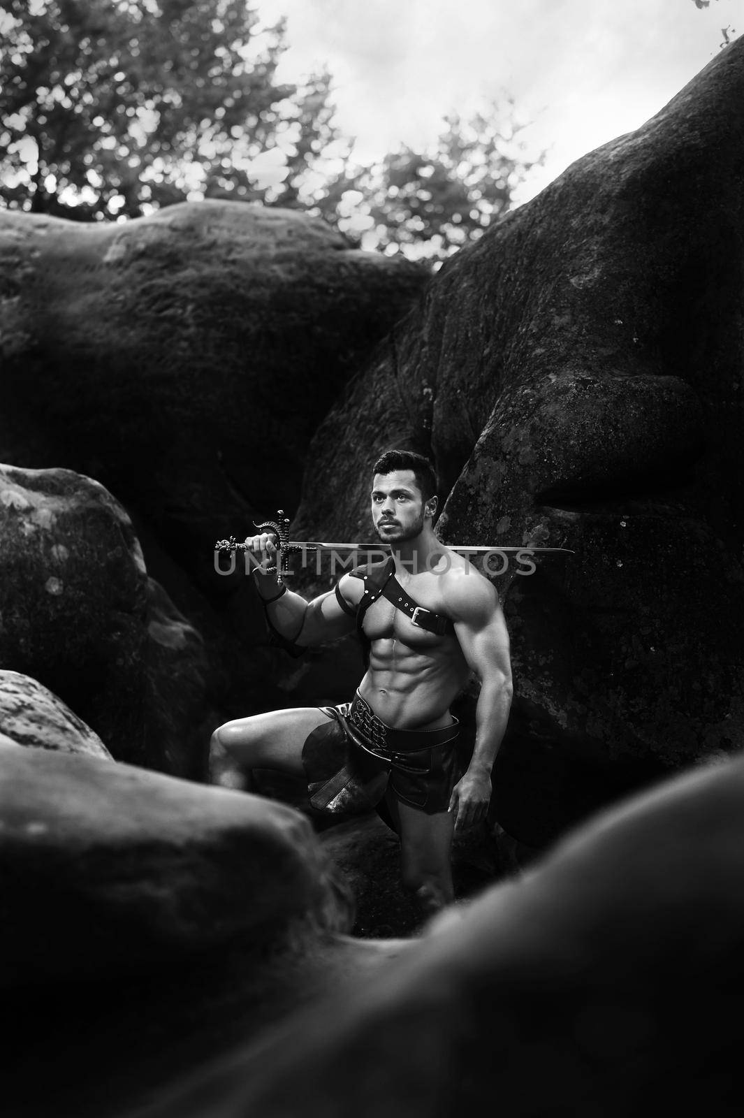 Soldier at heart. Monochrome shot of a young muscular man in a warrior outfit standing near the rocks holding the sword looking away