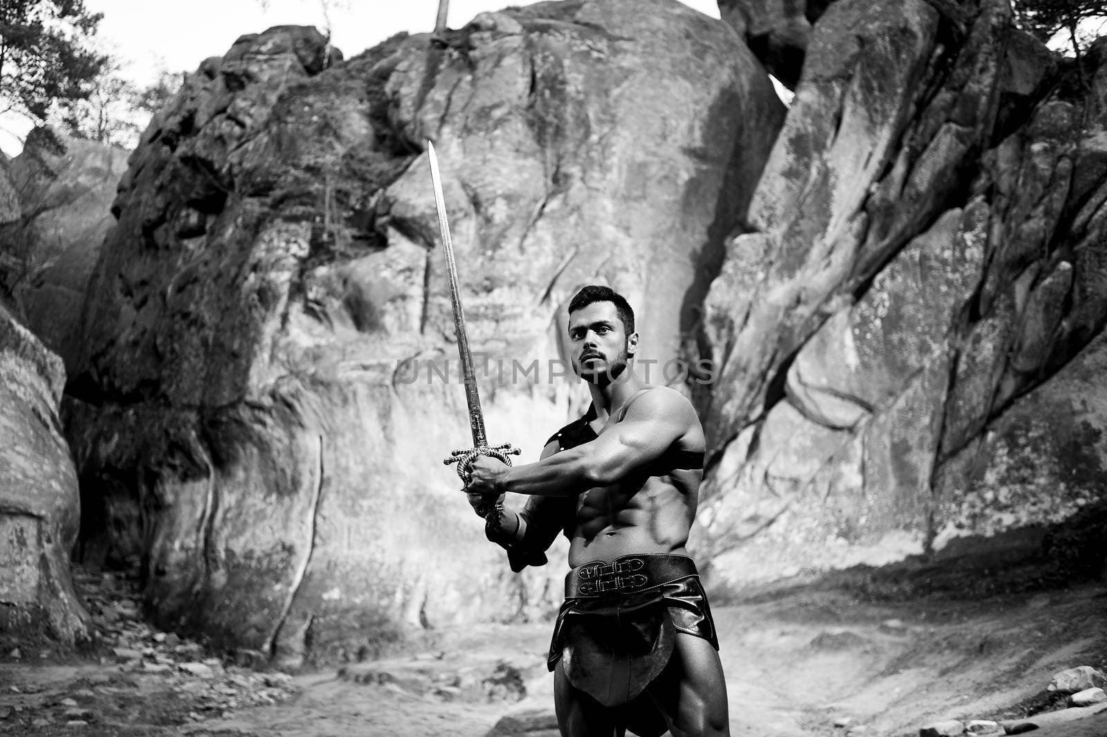 Getting ready for his battle. Monochrome portrait of a courageous manly warrior with a sword posing bravely near the rocks