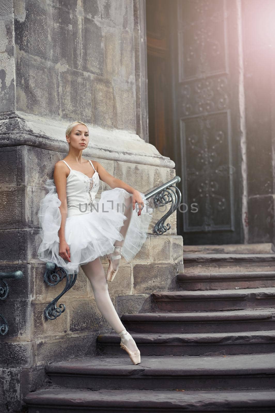 Portrait of a beautiful young blonde ballerina in white outfit standing gracefully on the stairs of an old castle.
