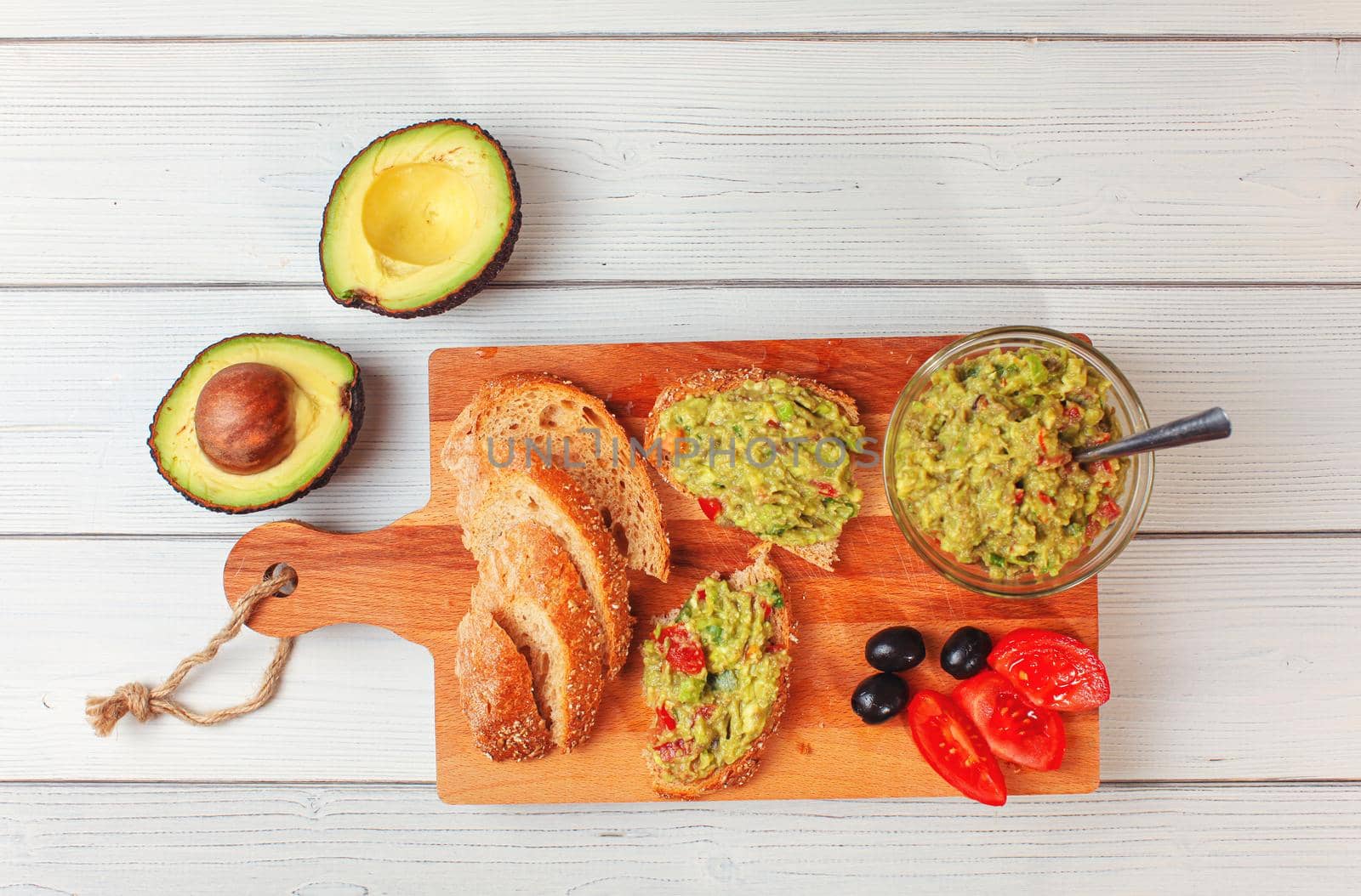 Flat lay photo, freshly prepared guacamole in small glass bowl, bread, tomatoes, olives at working board and two avocados next white wood desk.