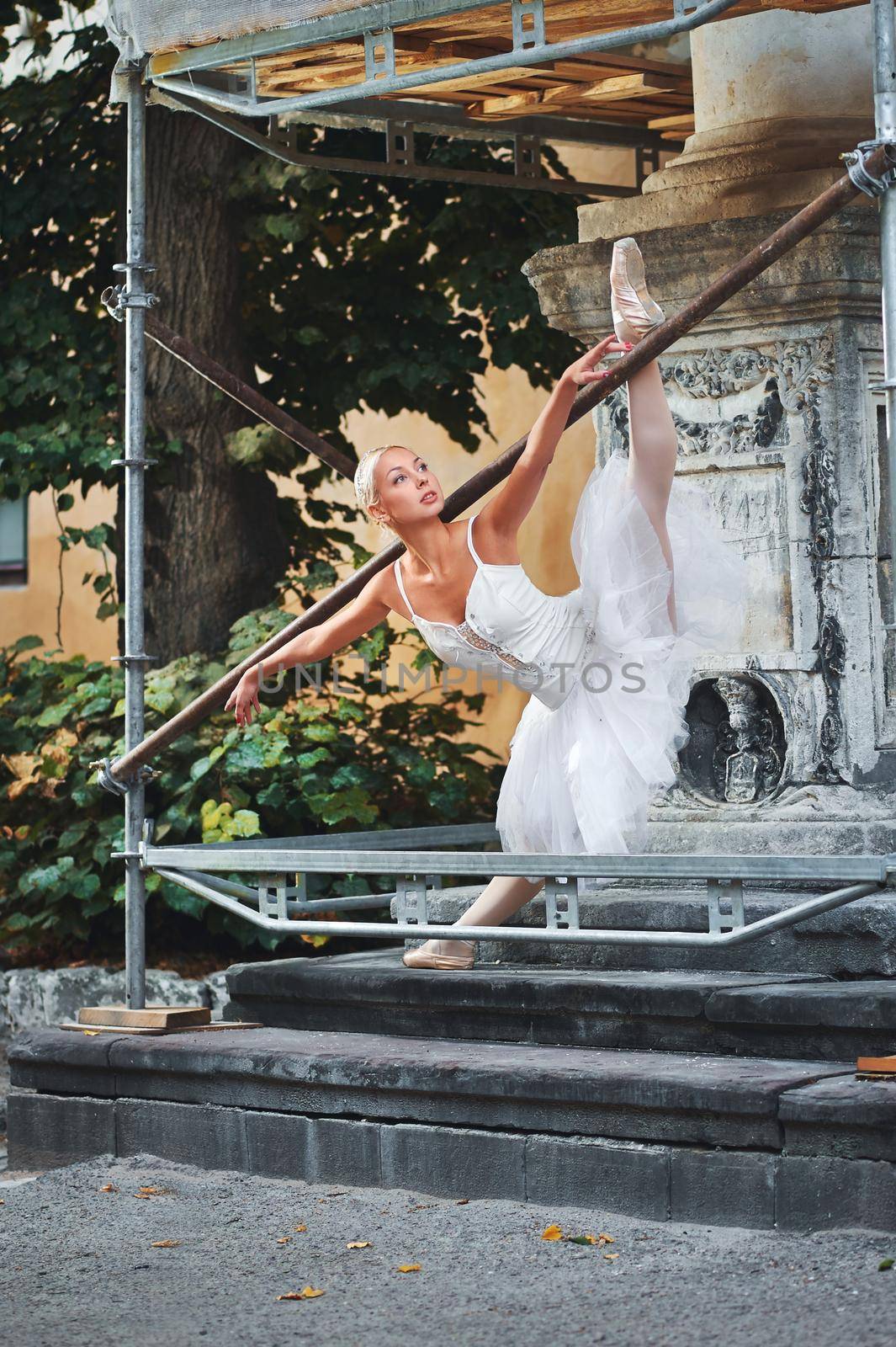 Vertical shot of a gorgeous ballerina in white outfit performing near an old building under construction.