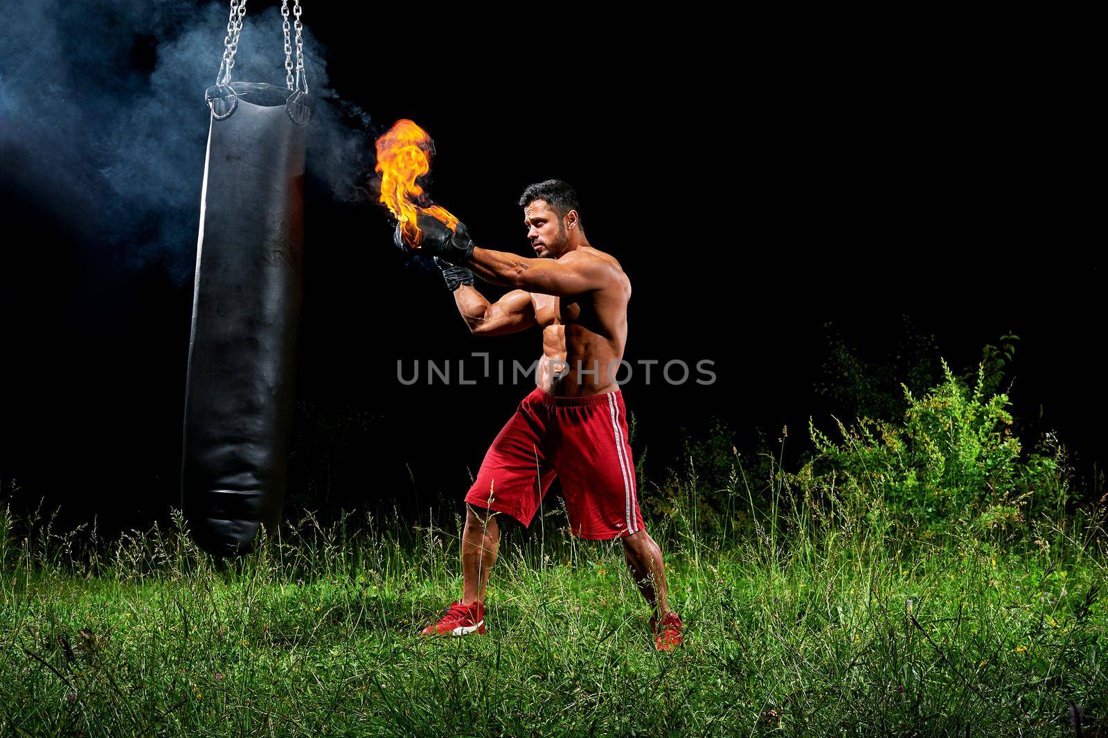 Sport motivation achievement concept. Professional male boxer training on a punching bag outdoors at night wearing burning boxing gloves. Professional martial arts fighter working out on punching bag