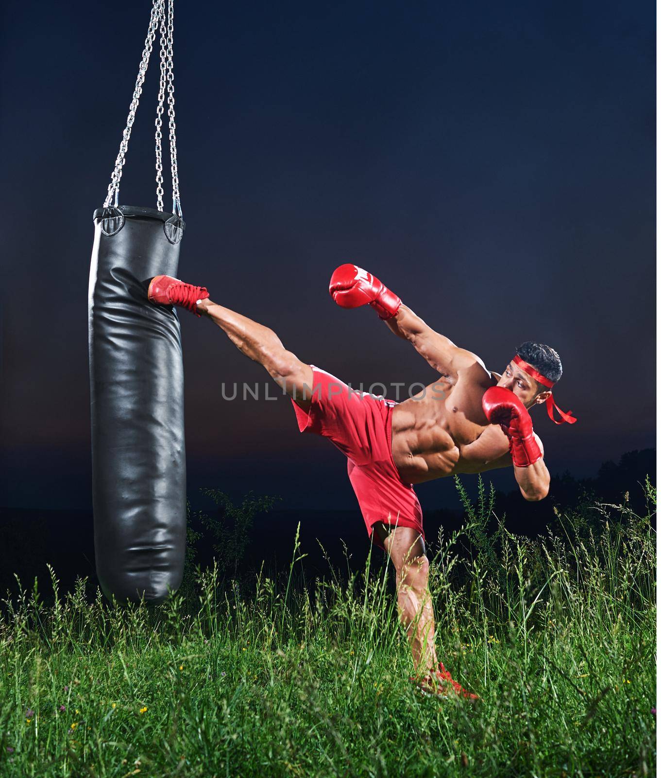 Handsome young strong muscular fighter exercising with a punching bag practicing his high kick martial arts combat fighting kickboxing Muay Thai strength power masculinity energy effort athlete.