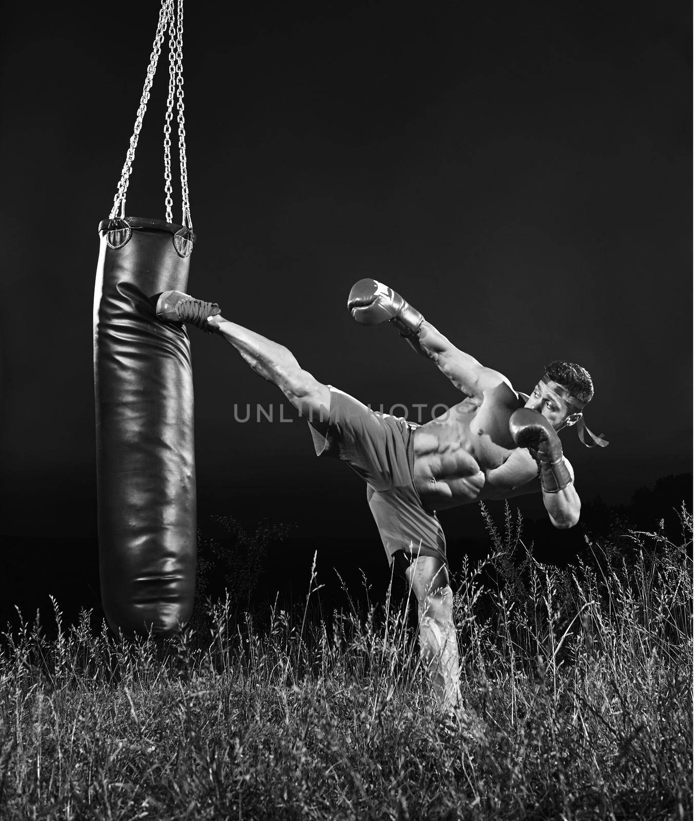 Full length monochrome shot of a muscular young kick boxer kicking a heavy bag training outdoors nature workout exercising combat martial fitness sport sportive sportsman athletics physique.