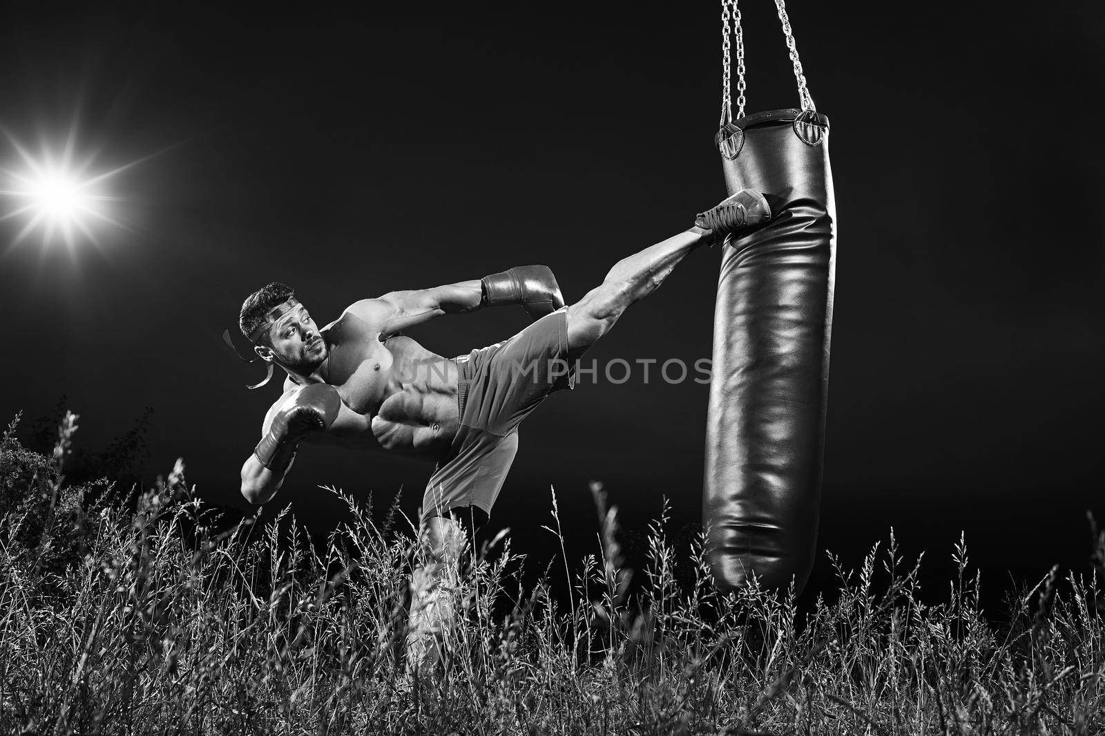 Black and white shot of a professional kick boxer practicing on a heavy punching bag outdoors at night copyspace motivation lifestyle workout muscles power strength masculinity martial sportsperson.