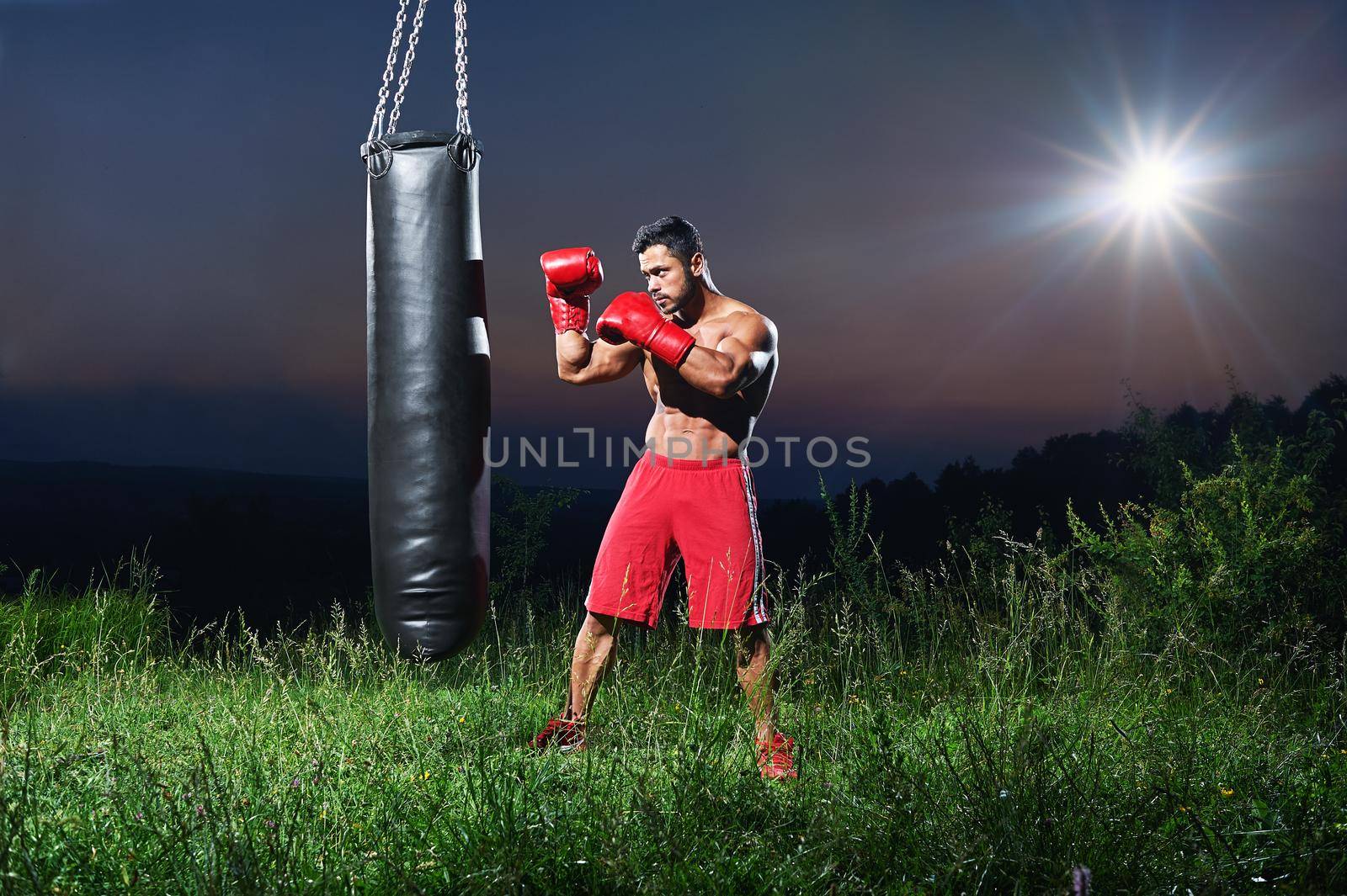 Full length shot of a muscular powerful boxer training shirtless outdoors at night working out with a punching bag copyspace nature concentration motivation sport athlete sportsman fighter combat.