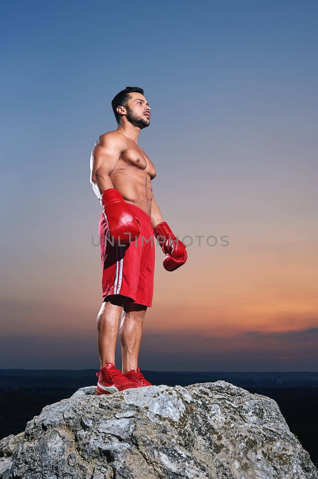 Low angle full length shot of a handsome young strong muscular male fighter wearing boxing gloves looking away standing outdoors on sunset peace calm fighting confidence masculinity fitness champion concept.