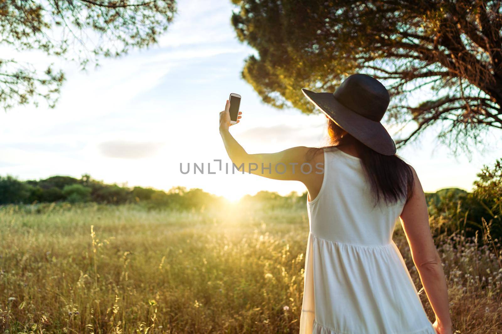 Suggestive scene from back of unrecognizable woman in white boho style dress and large dark hat taking a self portrait in front of the sun setting in the nature. Girl using cell for a self portrait