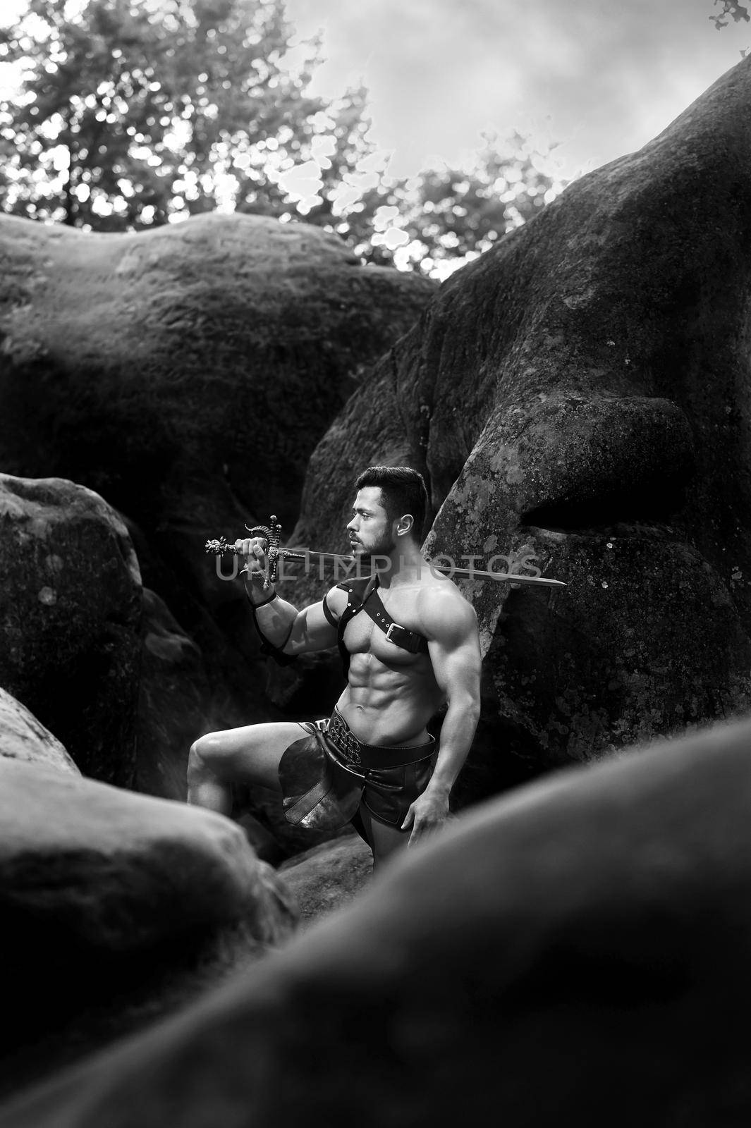 Forceful man. Monochrome vertical shot of a Spartan warrior resting near the rocks in the woods with a sword on his shoulder