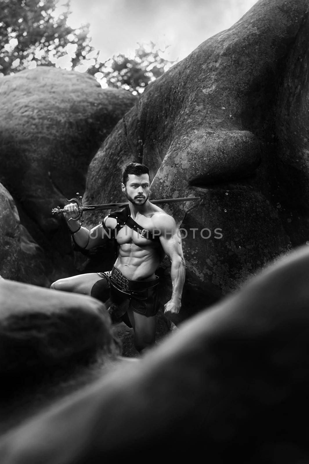 He knows no fear. Monochrome vertical portrait of a Spartan warrior with a sword looking around at the forest standing near the rocks