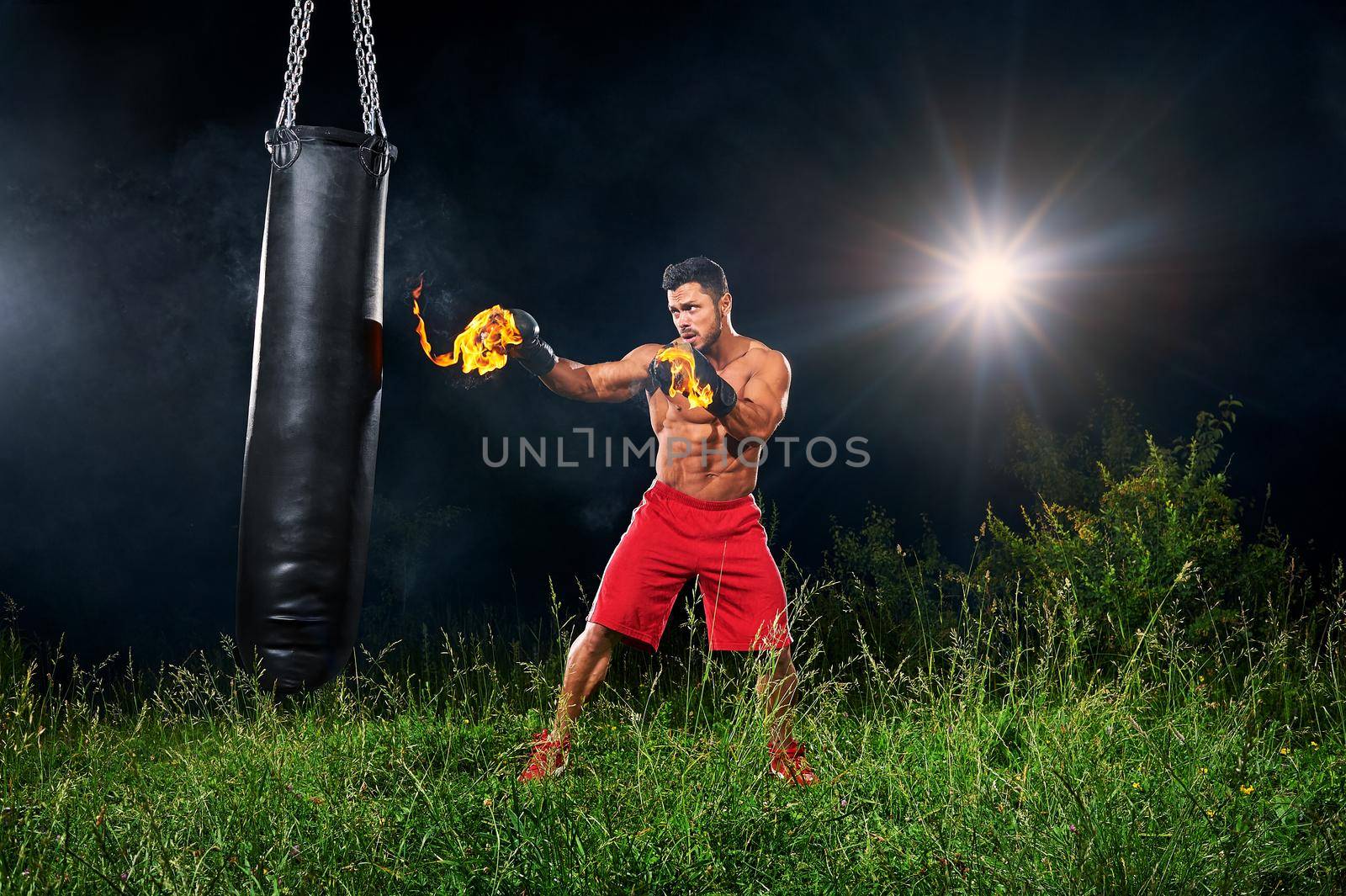 Full length shot of a muscular fit and toned young man boxing outdoors with burning boxing gloves training on a punching bag at night copyspace sports sportsman athlete athletics physique fitness.